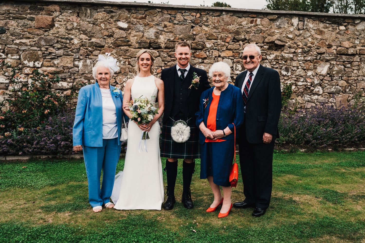 Wedding party poses for formal portrait against rustic stone wall.
