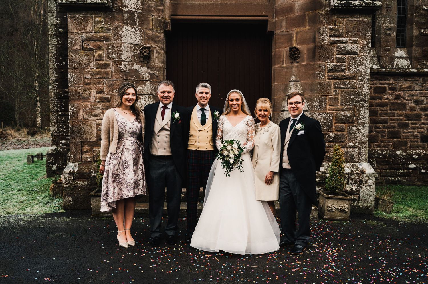 Small wedding group poses for a formal photo outside the stone church entrance.