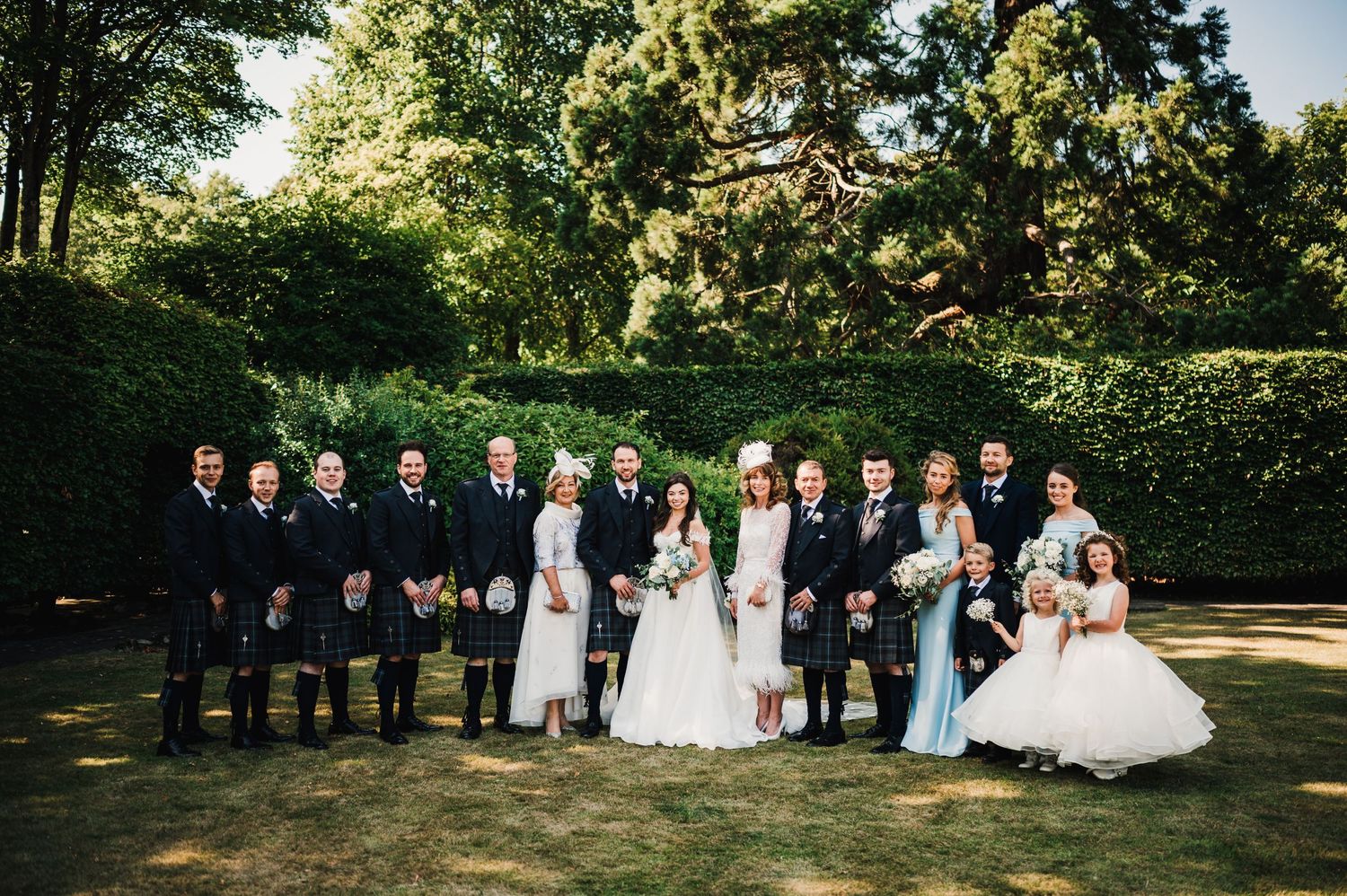 Formal wedding party line-up featuring Scottish kilts and elegant dresses in garden setting.