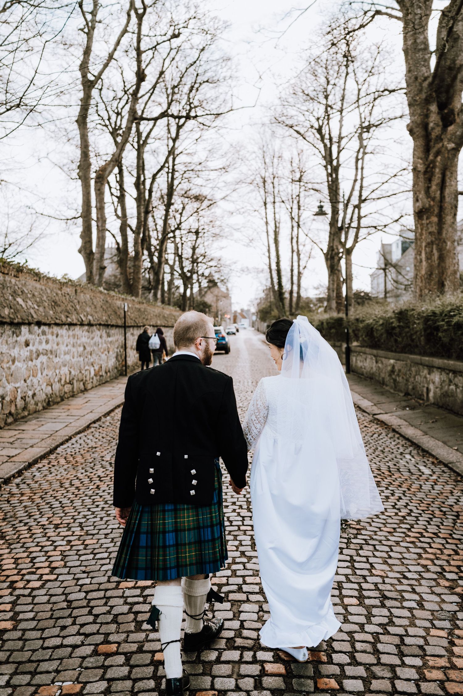 A wedding couple walks away on a tree-lined cobblestone path with stone walls on both sides.