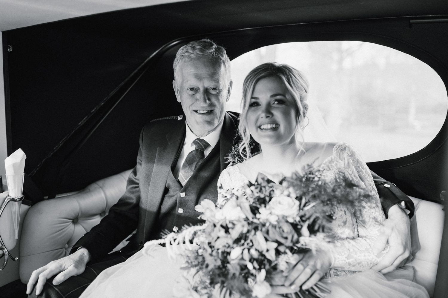 Black and white photo of couple in wedding attire sitting in classic car.