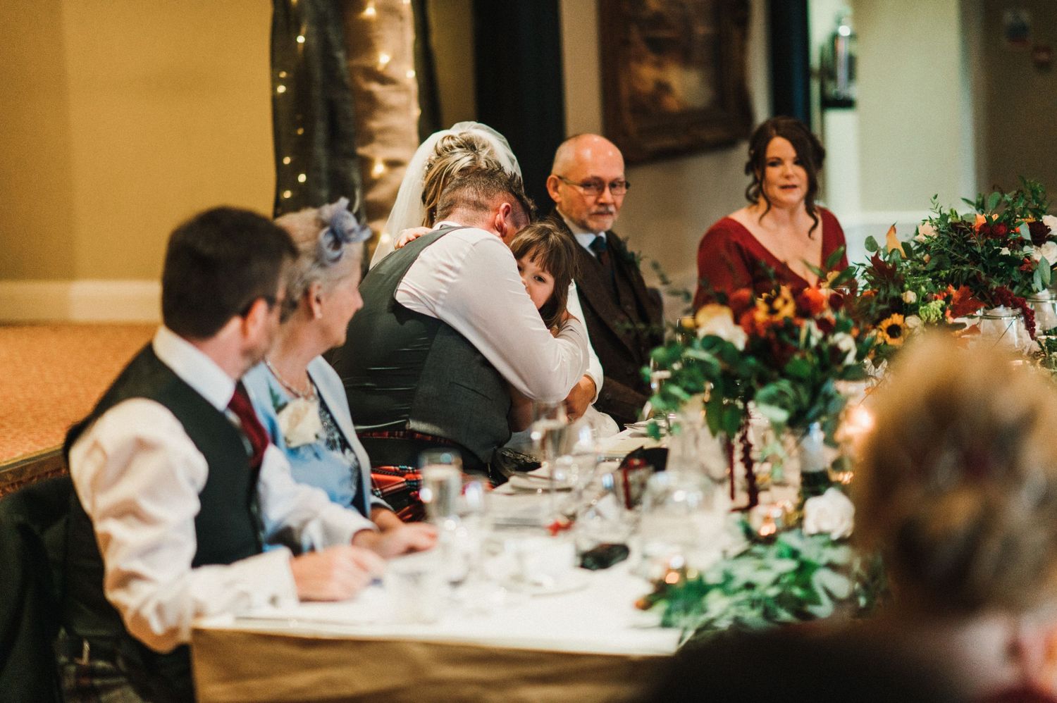 Wedding guests gathered around elegant tables decorated with greenery and flowers during an indoor reception.