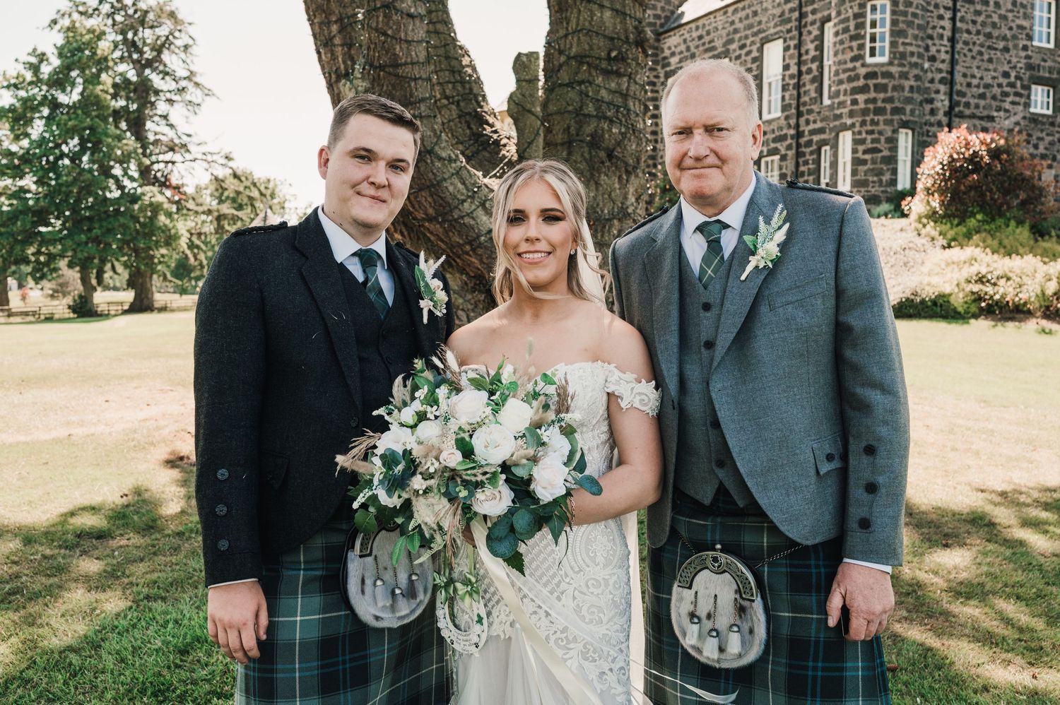 A wedding portrait with two men in kilts and a bride holding a white floral bouquet.