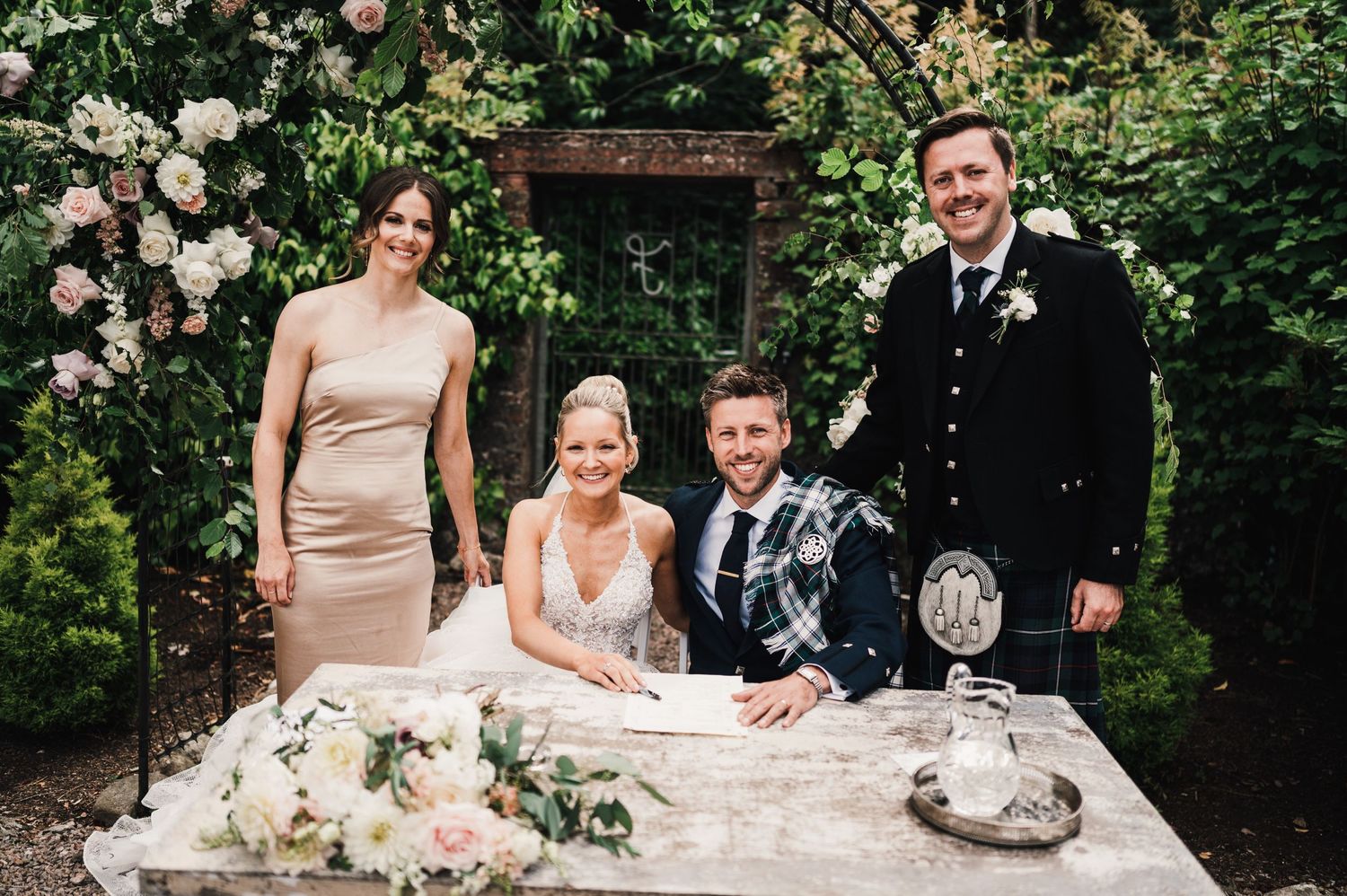 Wedding party gathered at stone table for document signing in garden setting with floral decor.