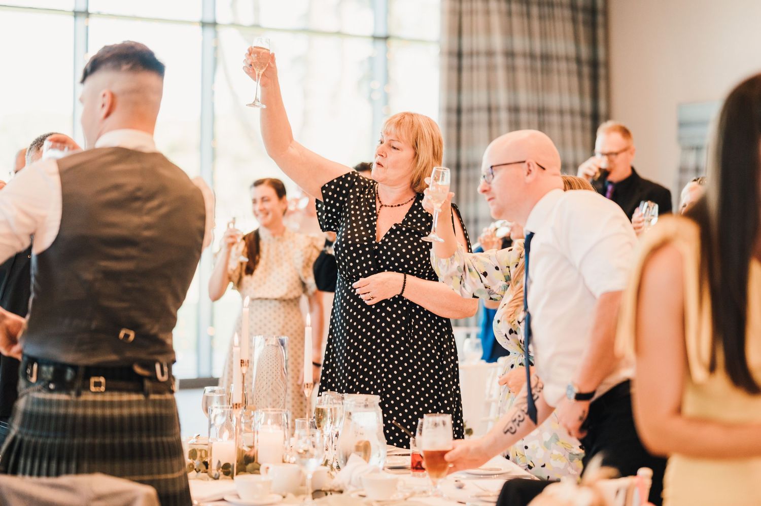 Wedding guests celebrate joyfully at a reception with people in kilts and polka dots dancing and cheering.