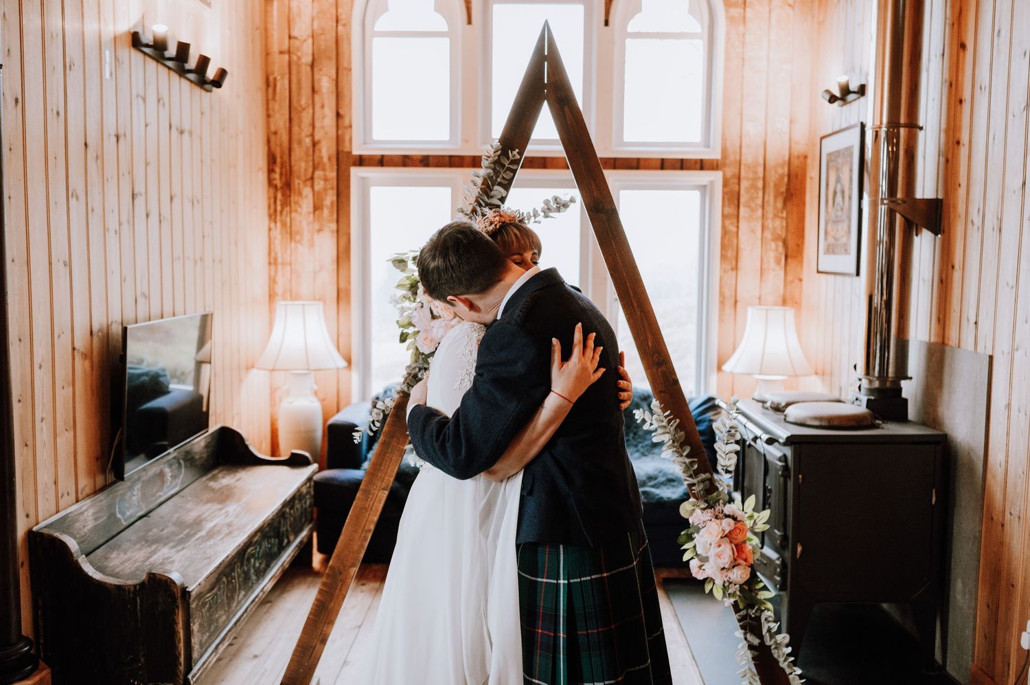 Intimate embrace between newlyweds in front of triangle-shaped altar with floral decorations.