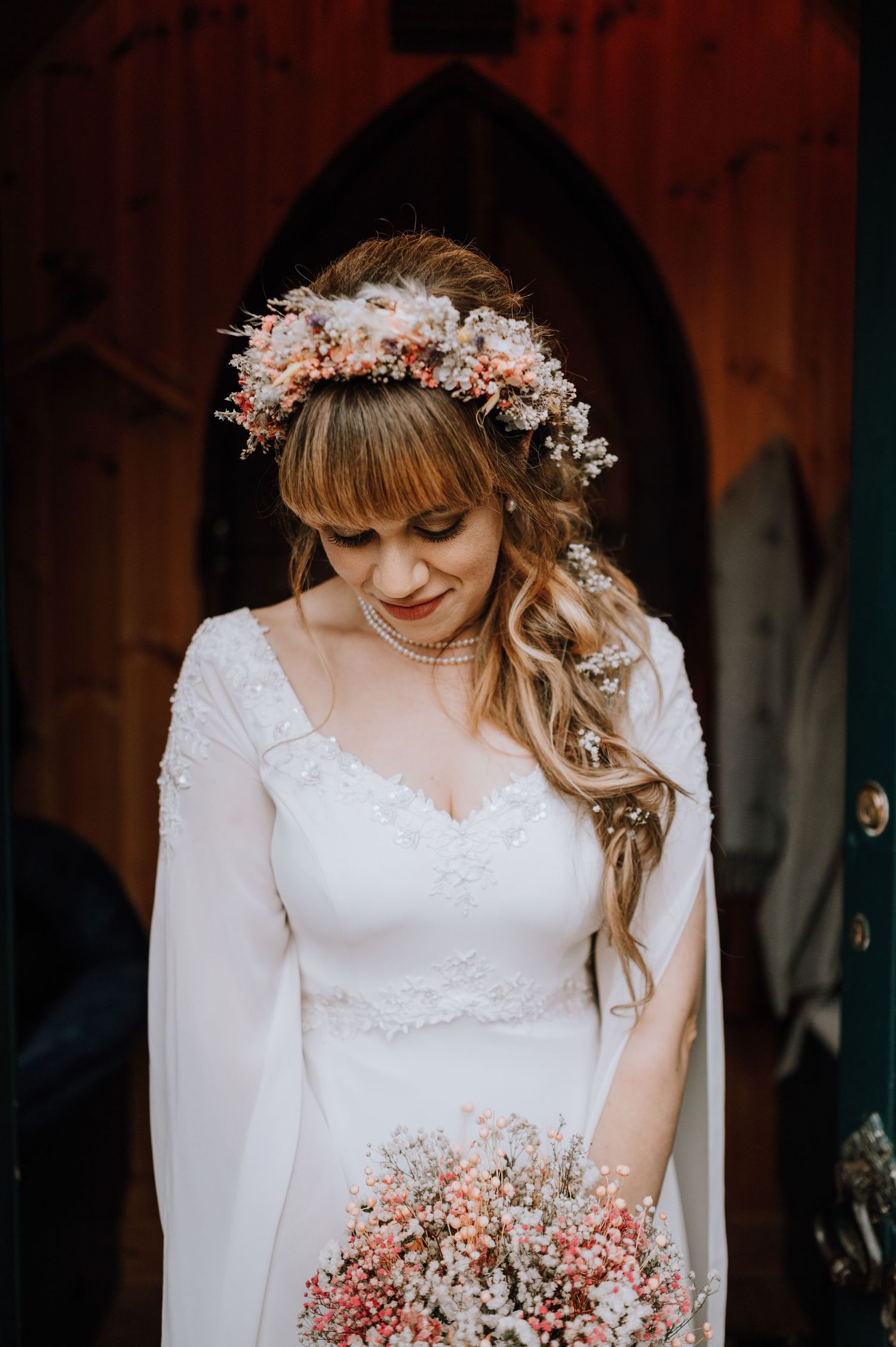 Bride wearing delicate floral crown and long sleeve white dress holds bouquet of pink flowers.