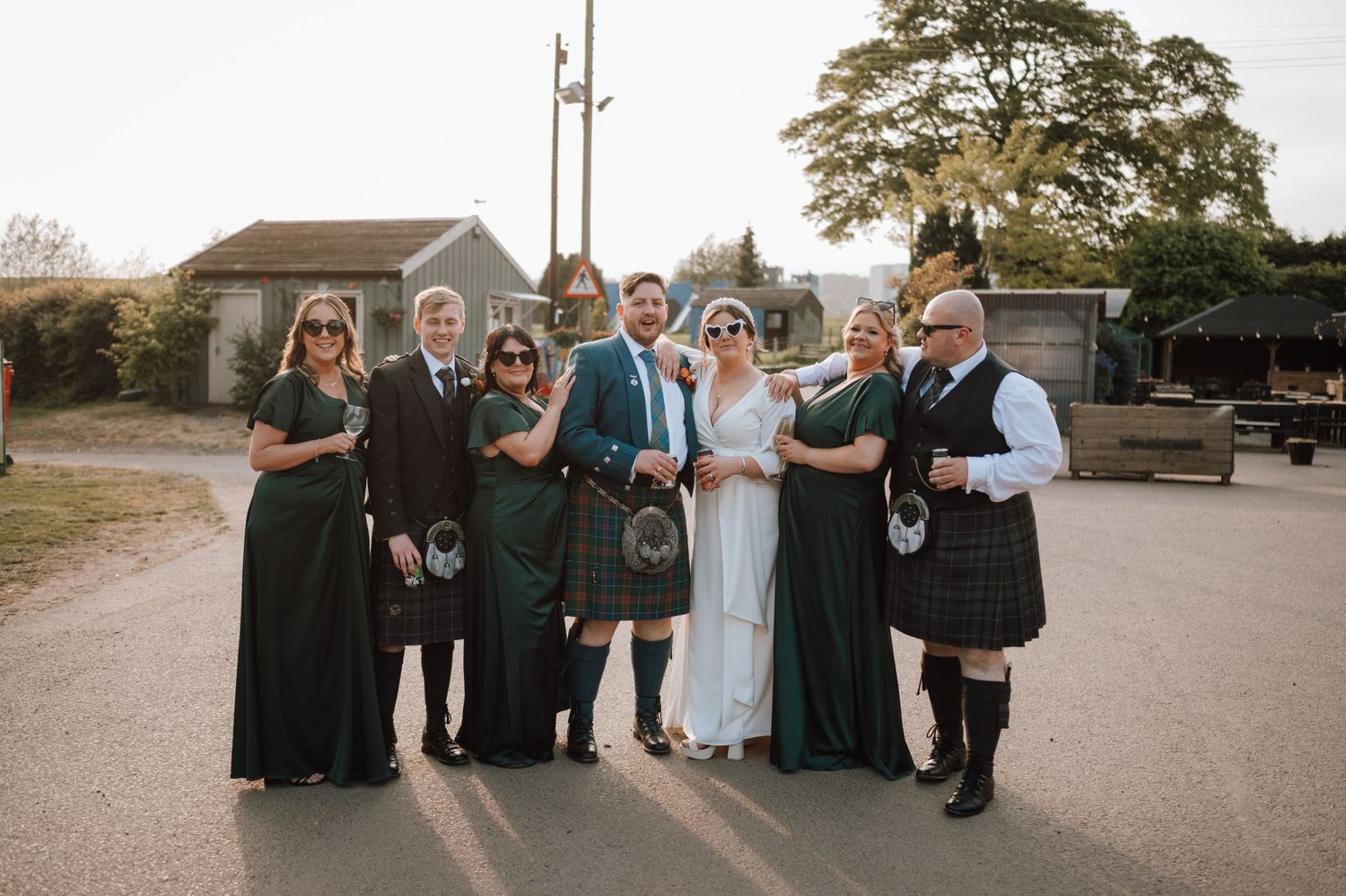 A formal group photo of a wedding party in Scottish attire poses on a country path.
