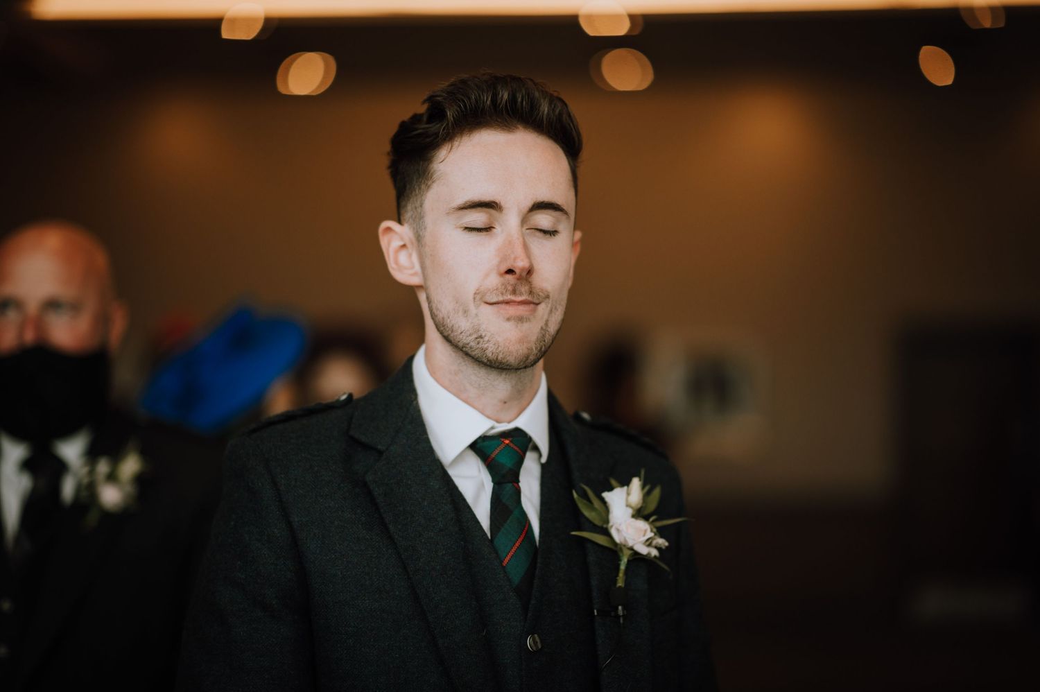 Wedding attendee in dark formal suit with boutonniere at indoor reception venue.