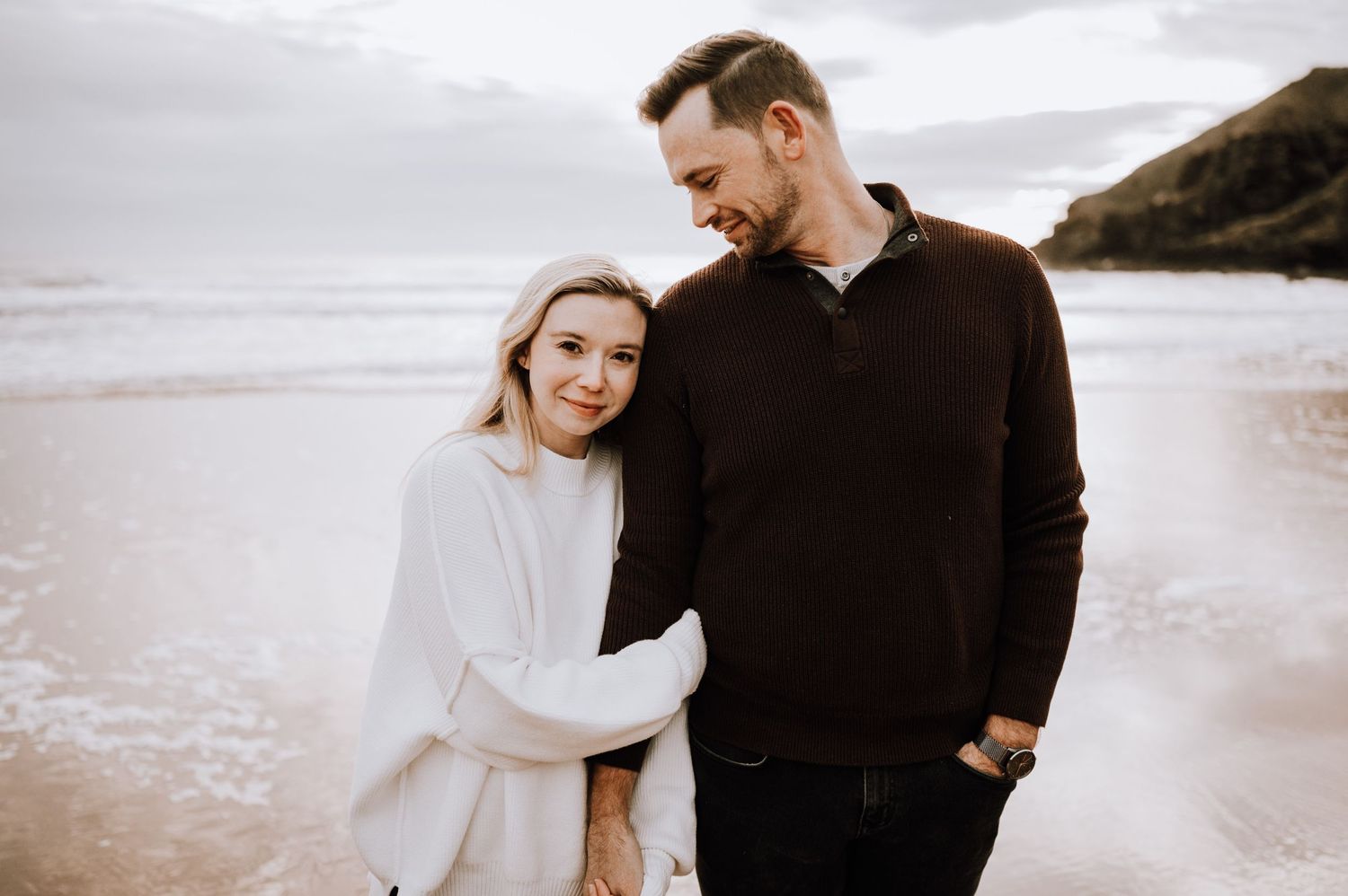 Couple in cozy sweaters sharing a sweet moment on an overcast beach.