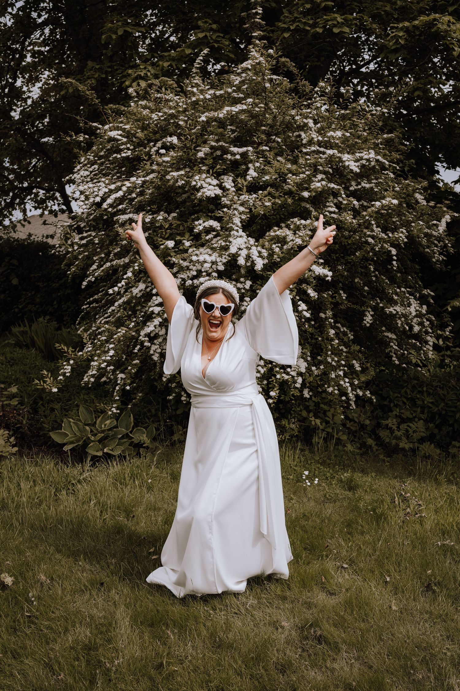 Bride in flowing white dress celebrates with arms raised in outdoor garden setting.