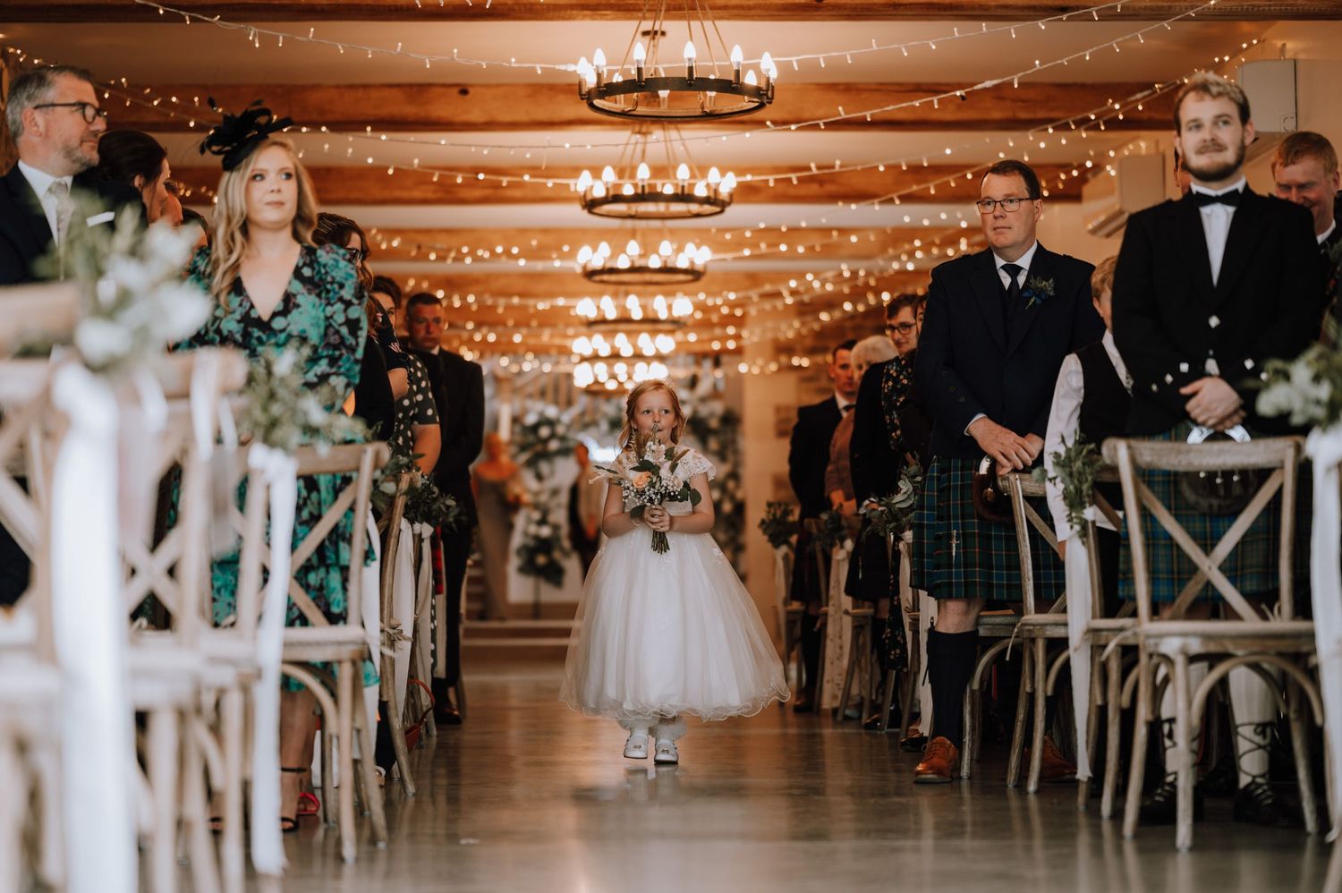 Young flower girl walks down aisle in elegant venue decorated with string lights and rustic wooden chairs.