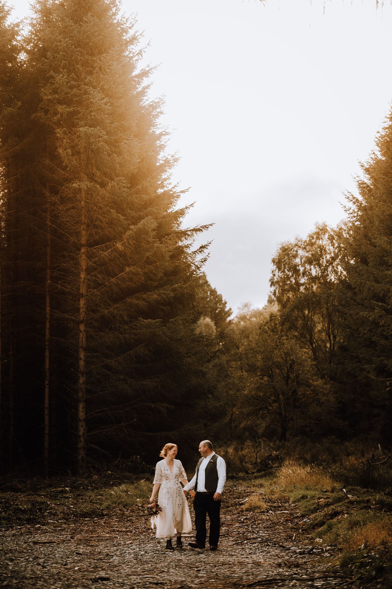 Autumn forest wedding scene with couple walking hand-in-hand down wooded path.