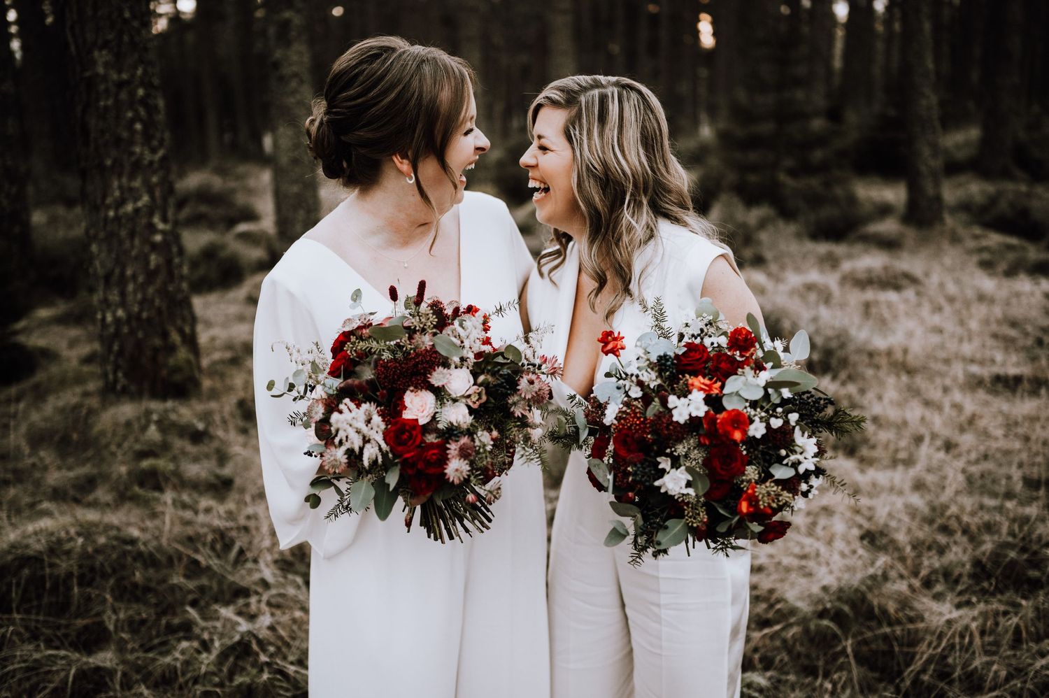 Same Sex Wedding Photography. Two figures in flowing white dresses share an intimate moment among tall pine trees at sunset.