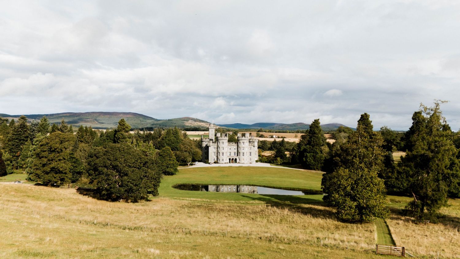 Wide panoramic view of a castle estate nestled among green hills and woodland.