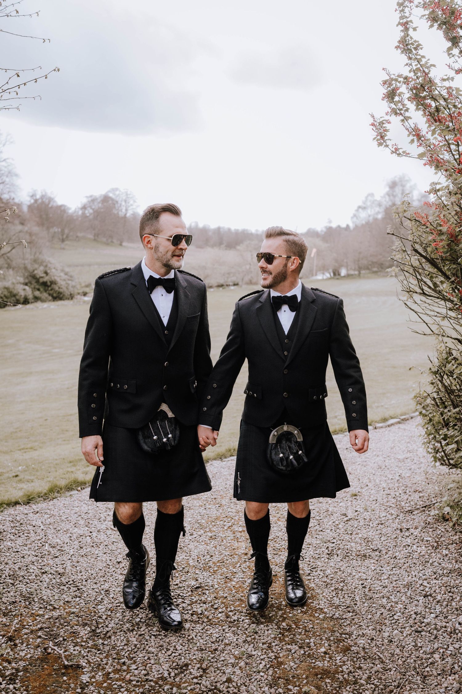Logie House Wedding Photography. Two people in matching black tuxedos walk hand in hand on a gravel path.