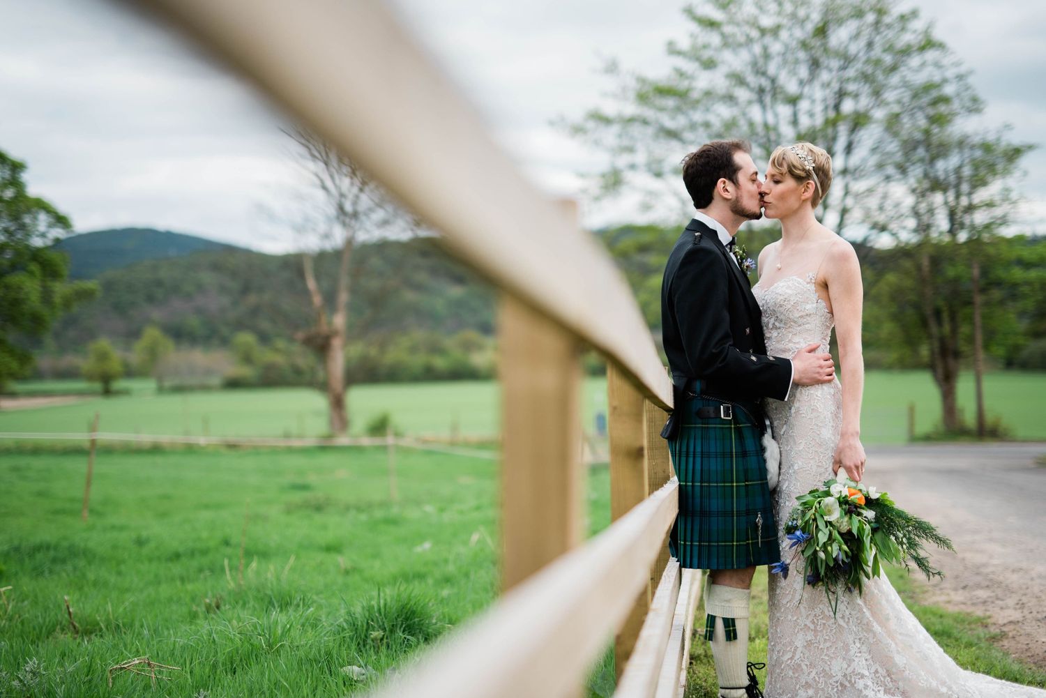 A romantic countryside wedding photo with the couple standing by a wooden fence in Scottish Highland scenery.