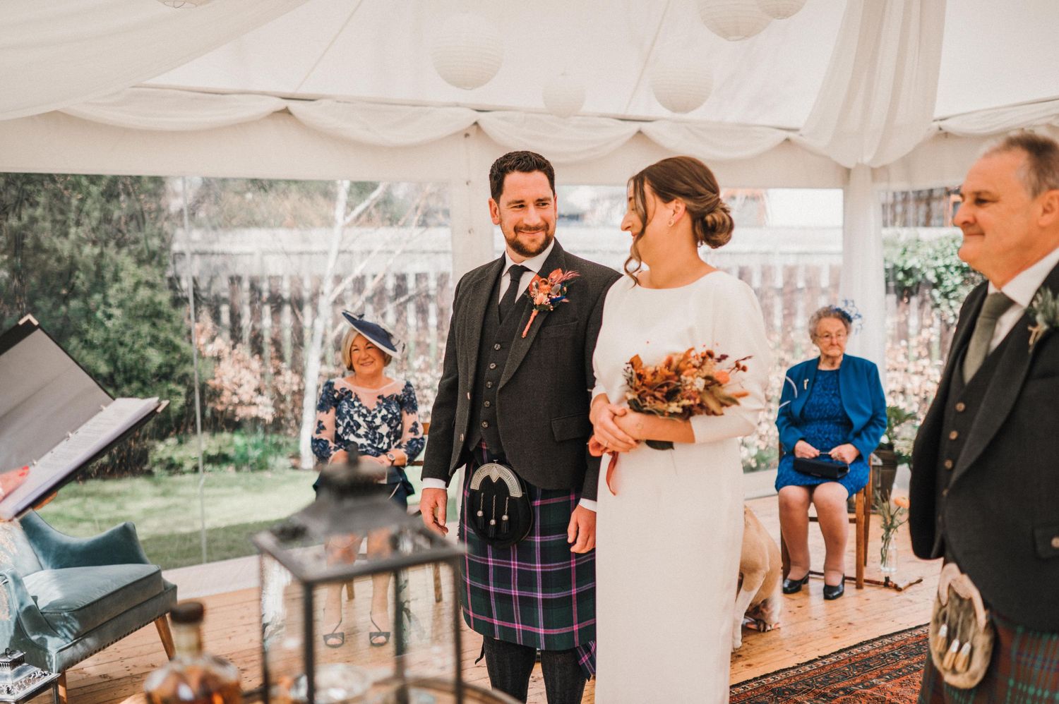A wedding ceremony takes place under a white tent with Scottish traditional elements.