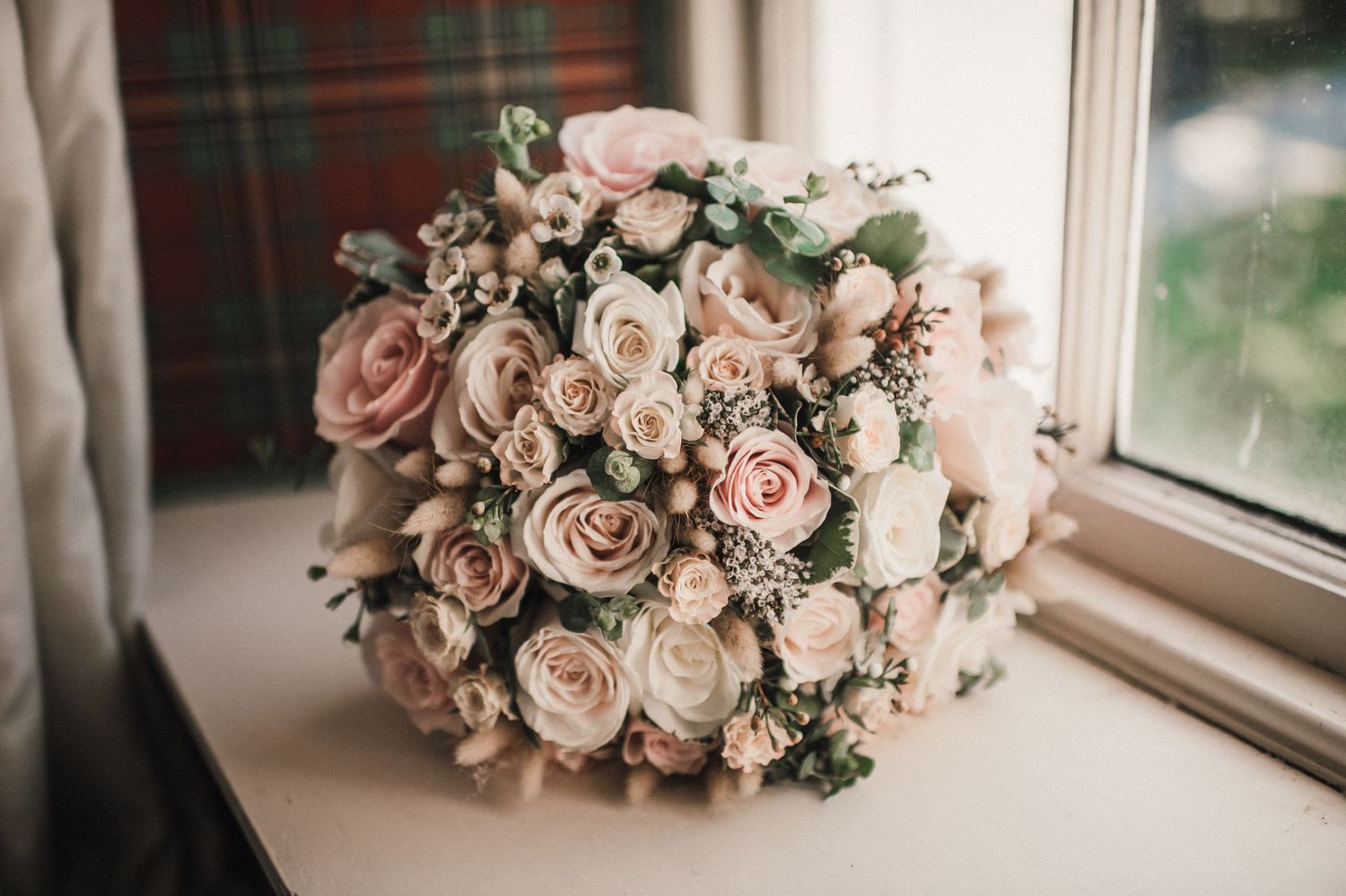 A stunning wedding bouquet with blush pink roses and delicate greenery by a window.