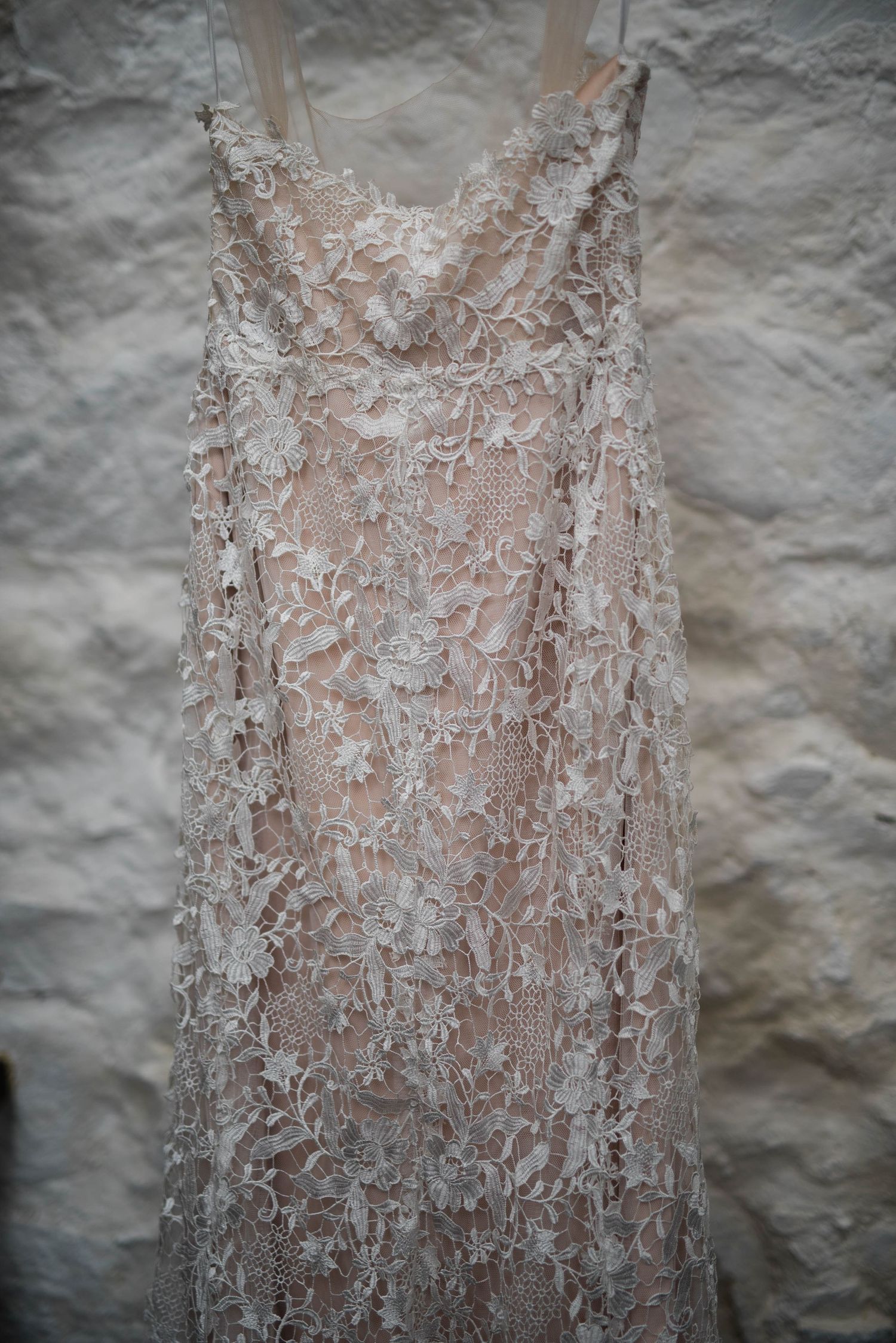 A delicate ivory lace wedding dress with intricate floral patterns displayed against a textured white wall.