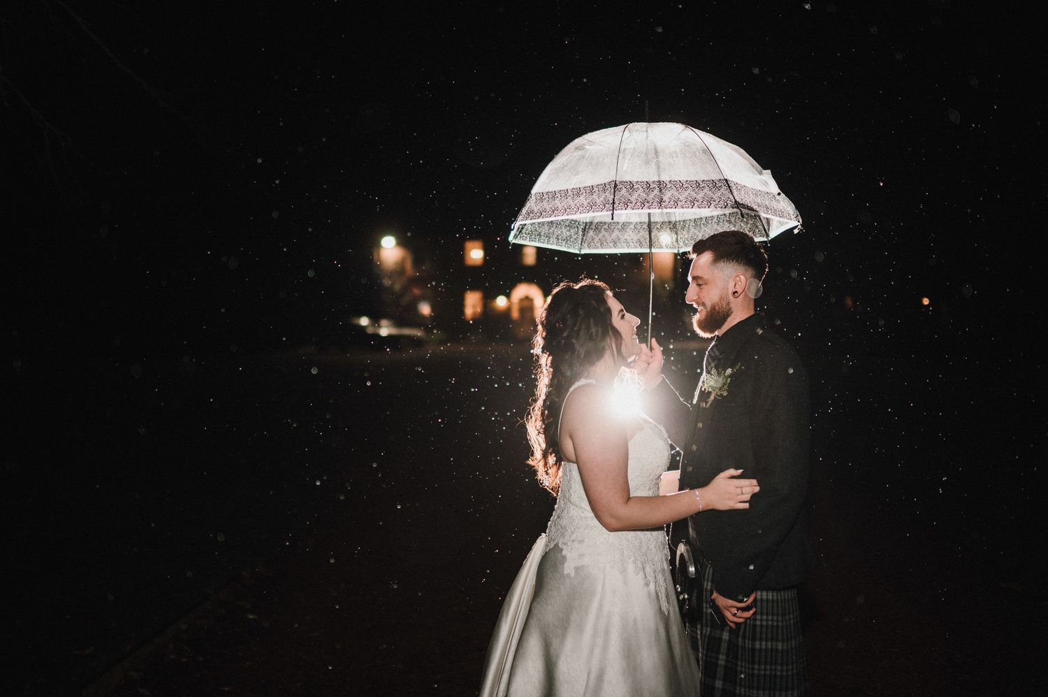 Couple shares romantic moment under clear umbrella at night with backlit rain.