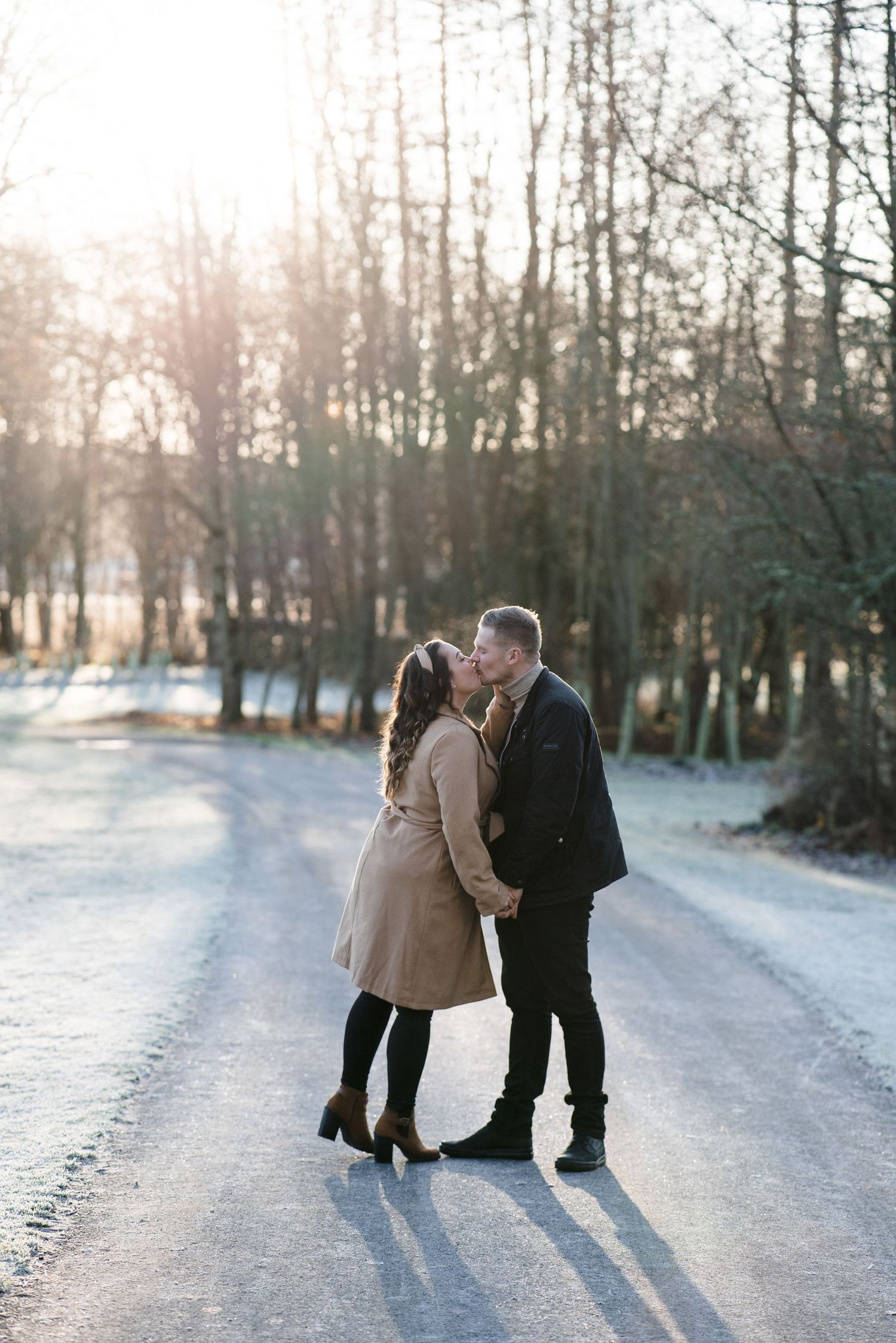 Romantic winter portrait of couple kissing on snow-covered path at sunset.