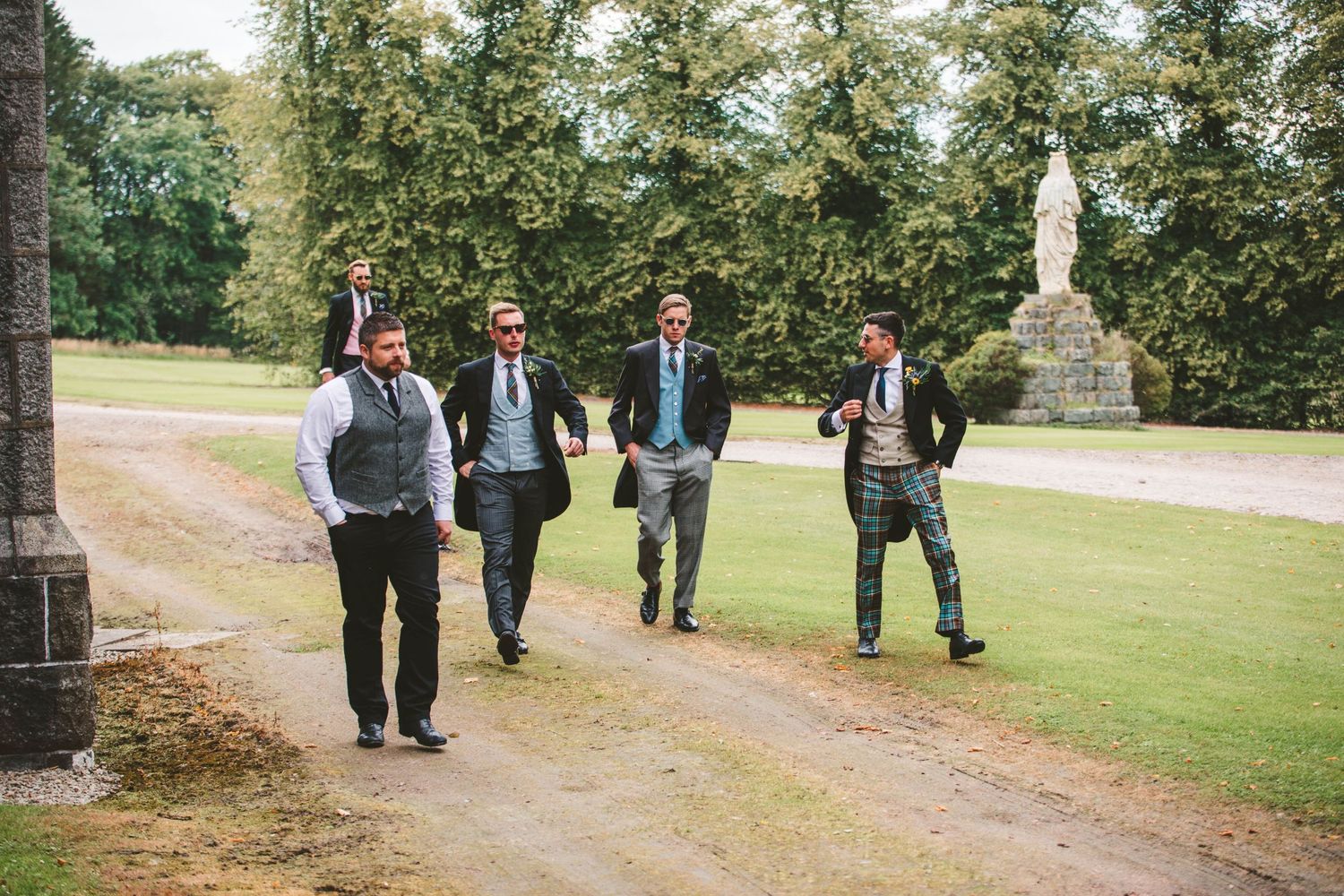 A group of well-dressed guests walk along a garden path at an outdoor wedding venue.