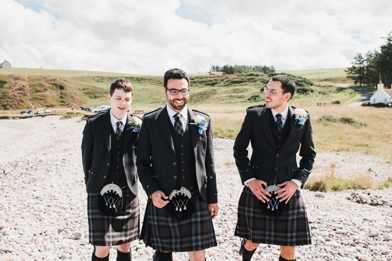Three men in formal kilts and jackets share a laugh on a gravel path.