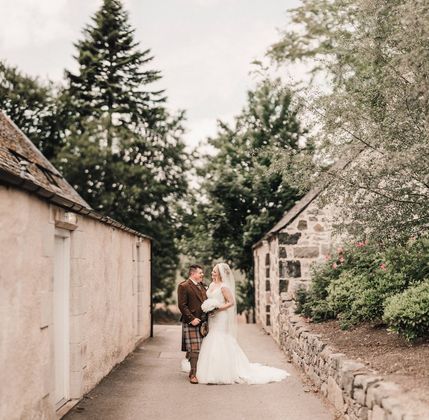 A wedding couple walks between stone walls along a rustic garden pathway.