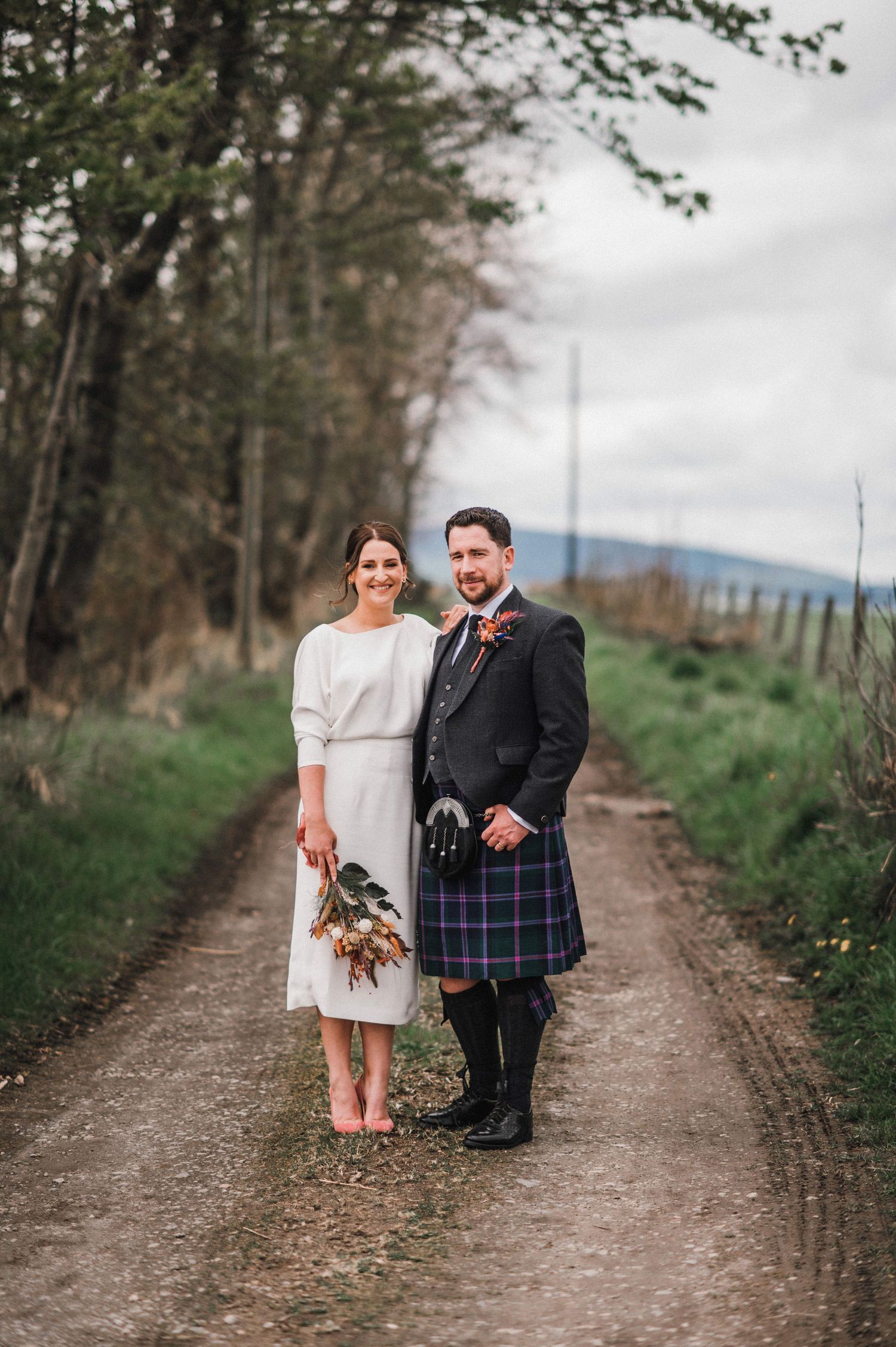 A countryside wedding portrait on a dirt path surrounded by trees with mountains in the distance.