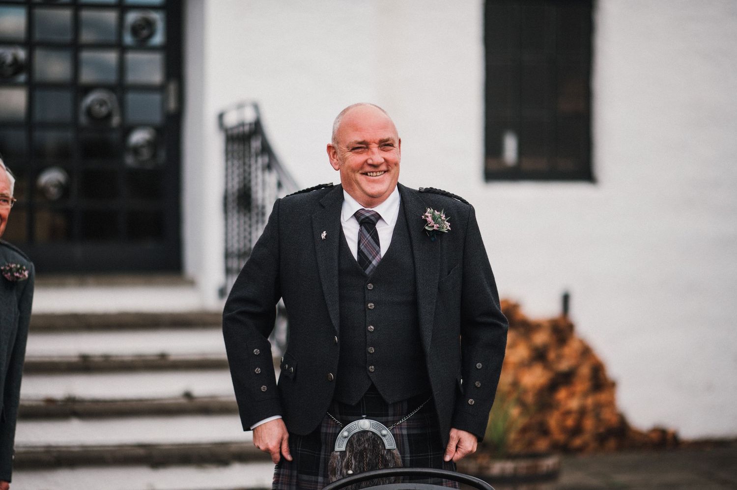A person in formal black attire and kilt stands smiling on white building steps.