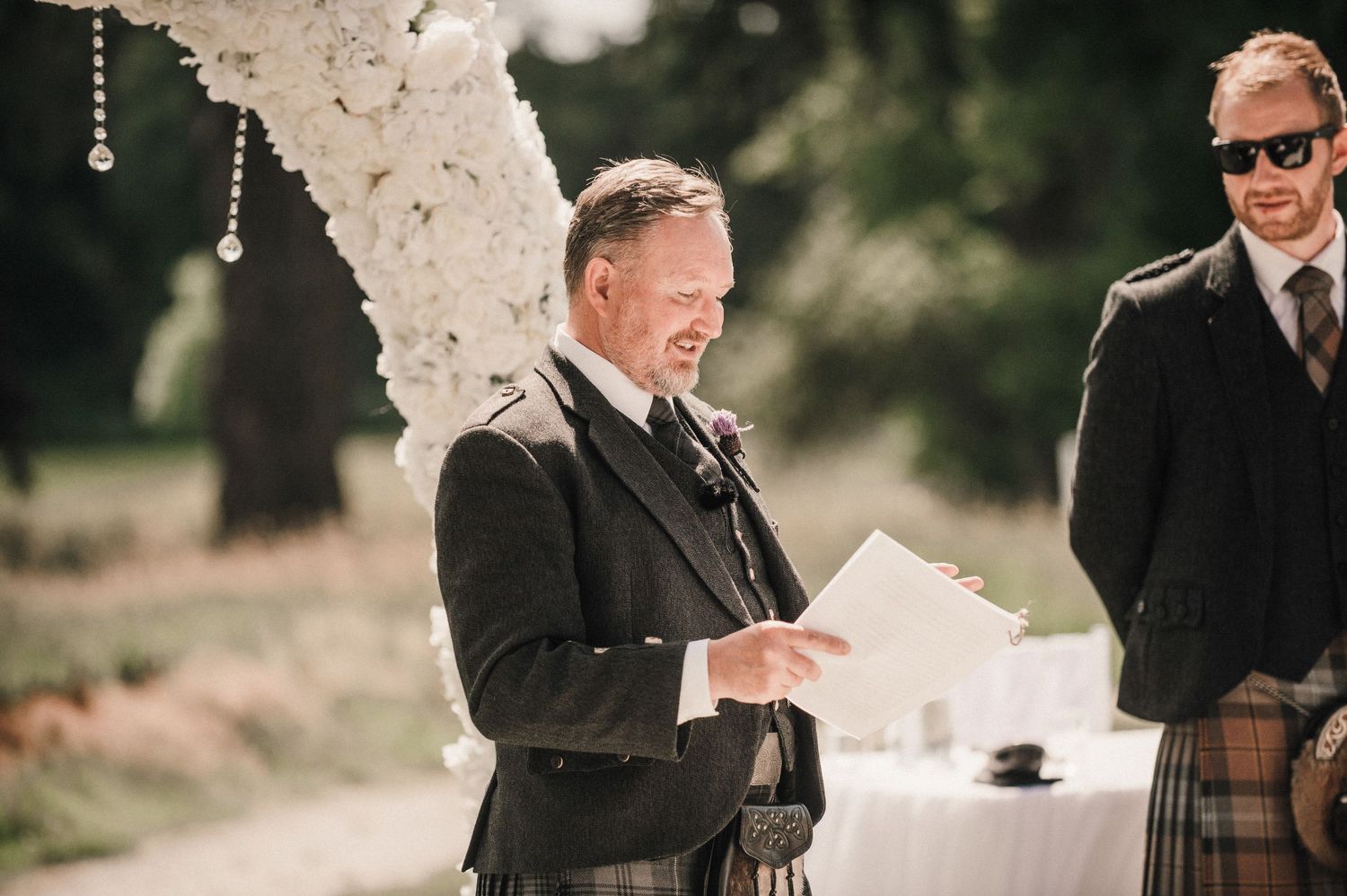 A person in formal attire reads from papers during an outdoor wedding ceremony.