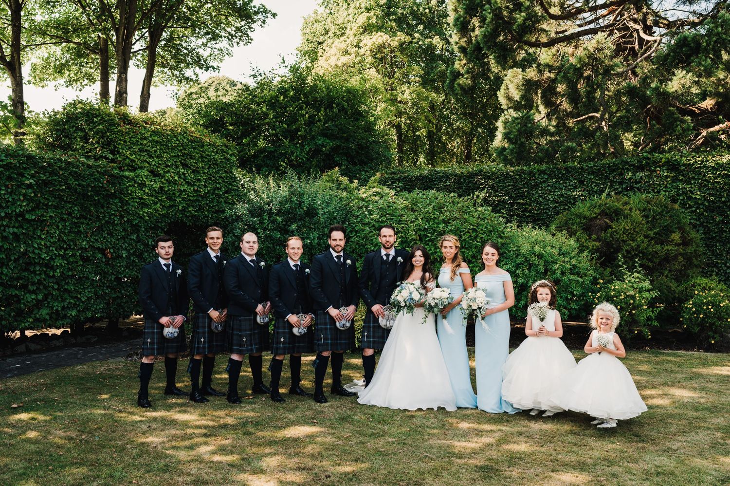 Wedding party poses on grass with groomsmen in kilts and bridesmaids in formal attire.