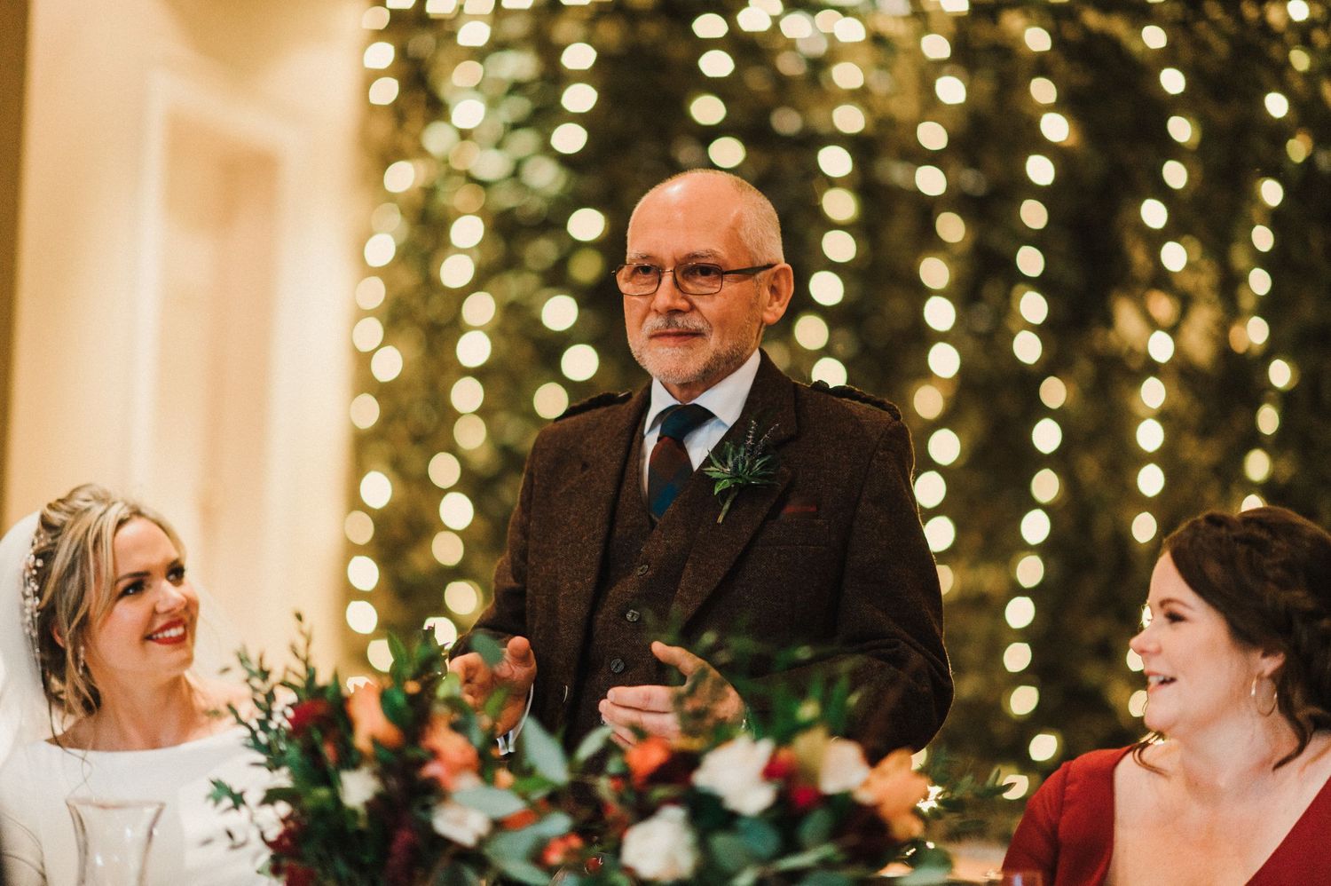 A celebratory moment during wedding speeches with twinkle lights and floral decorations in the background.