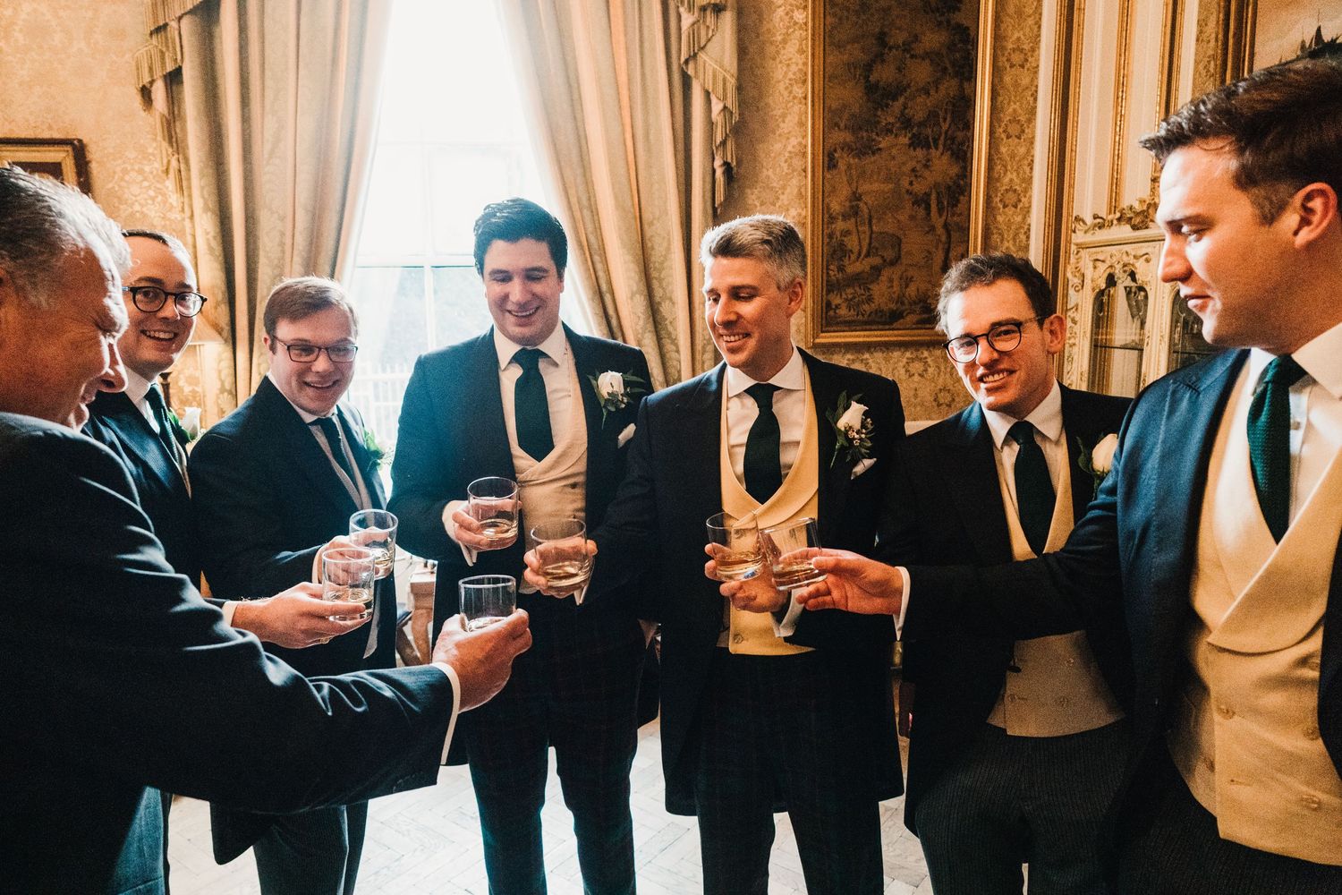 Group of groomsmen in dark suits toasting with drinks in an elegant room with tall windows.