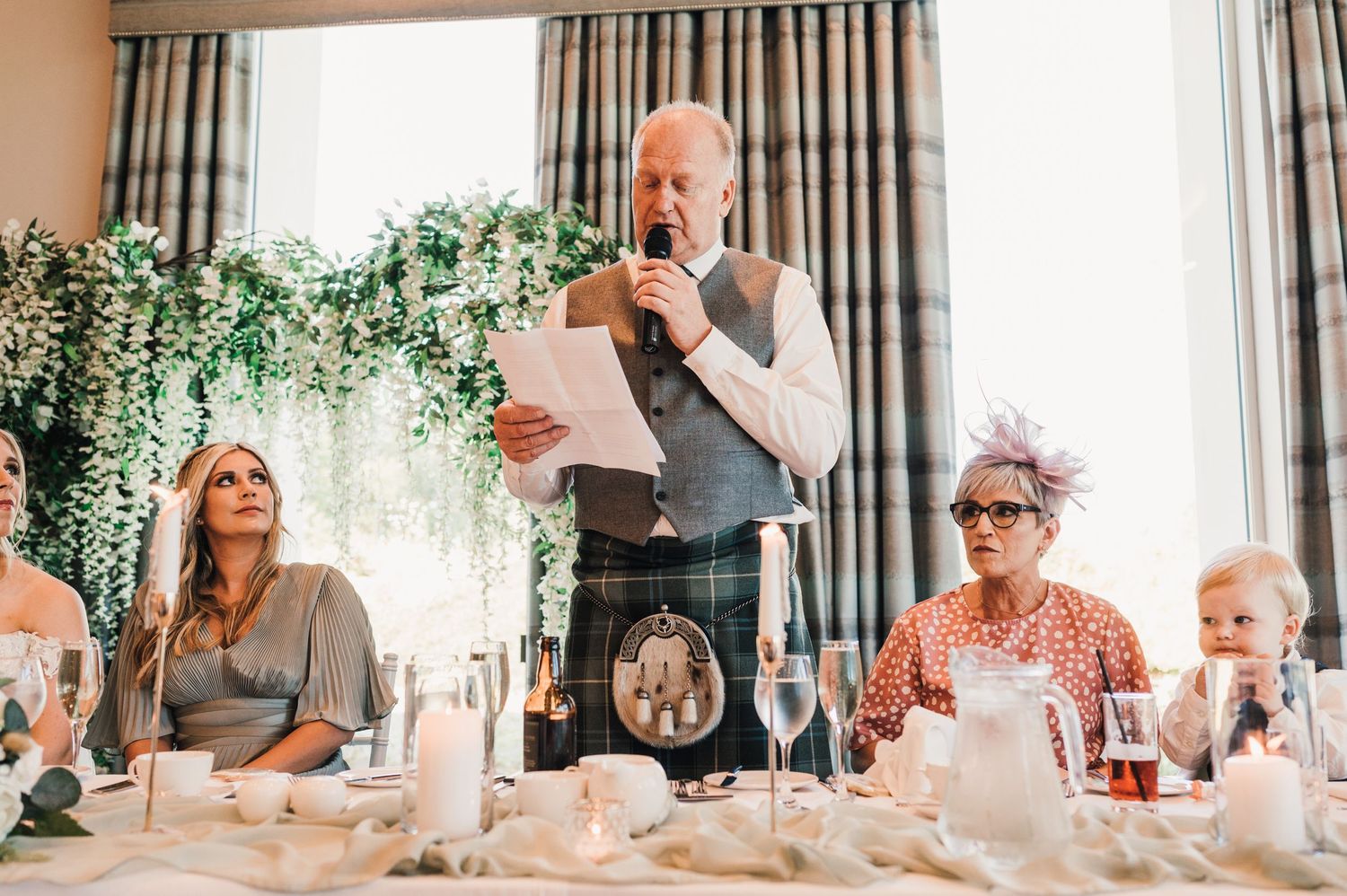 Speaker addressing wedding guests during reception dinner under hanging floral decorations.