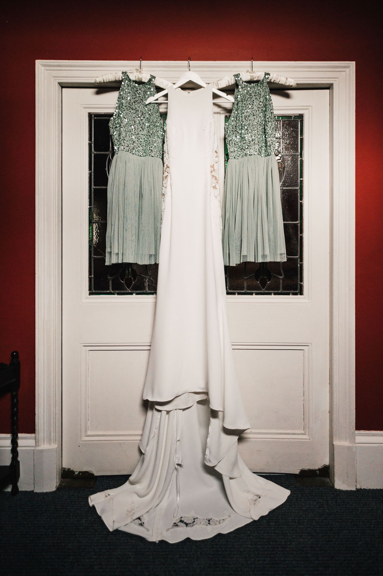 A white wedding dress hangs between two mint green bridesmaid dresses against white double doors.