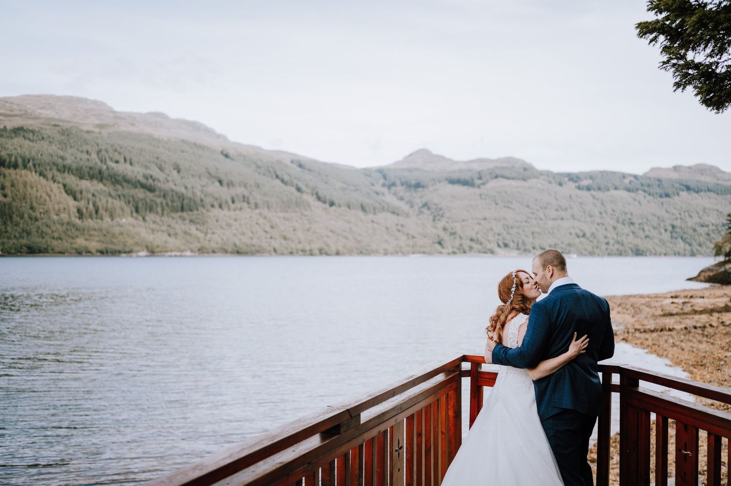 Couple embraces on wooden deck overlooking peaceful mountain lake during wedding photoshoot.