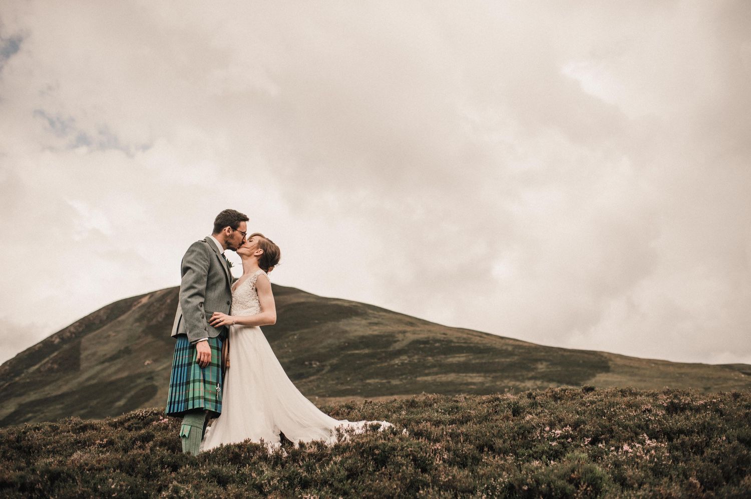 Wedding couple in Scottish Highland dress sharing an intimate moment atop a misty mountain.