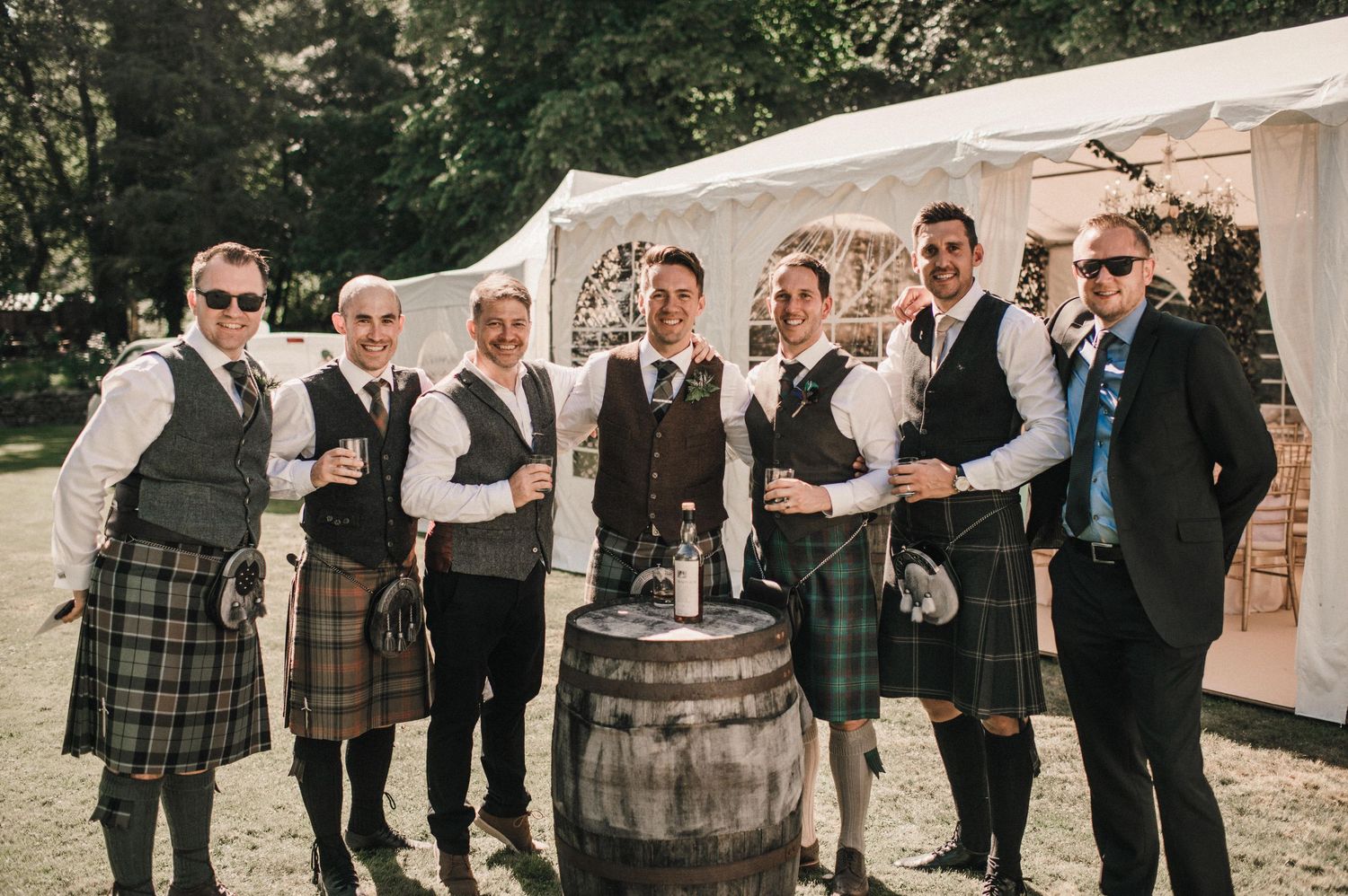 Groomsmen in kilts and waistcoats posing together with a rustic wine barrel outdoors.