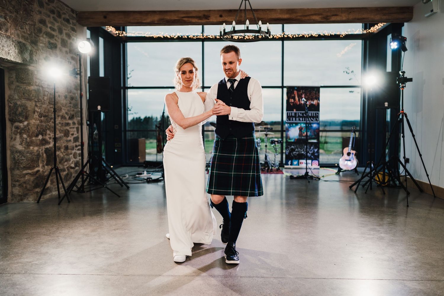Couple in traditional Scottish wedding attire dancing in a modern venue with large windows.