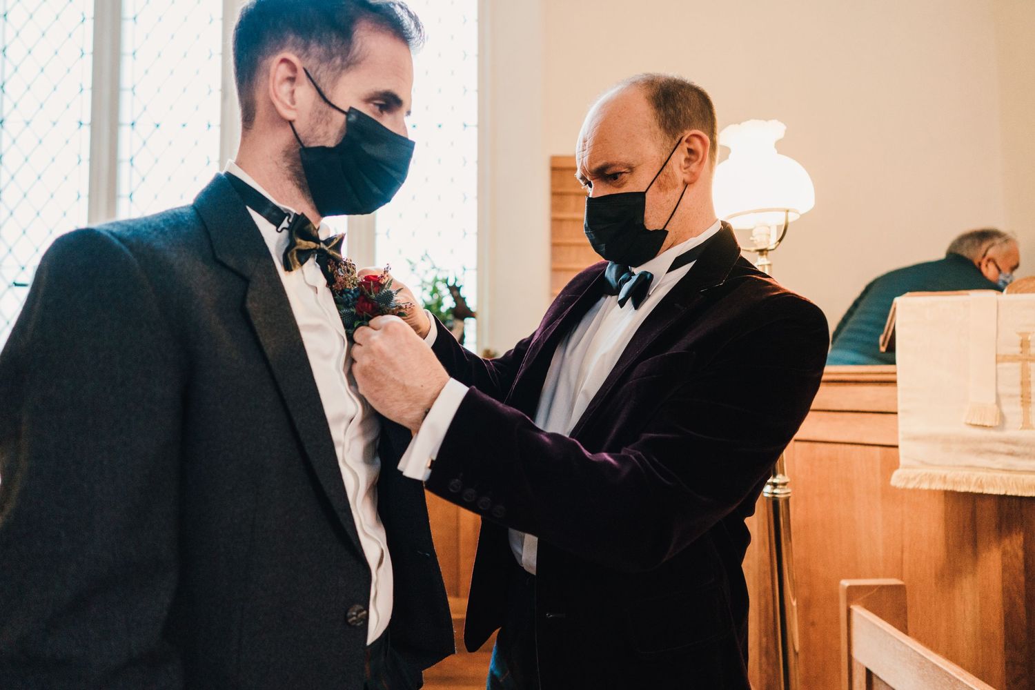 Two people in formal attire with face masks adjusting boutonniere before ceremony.