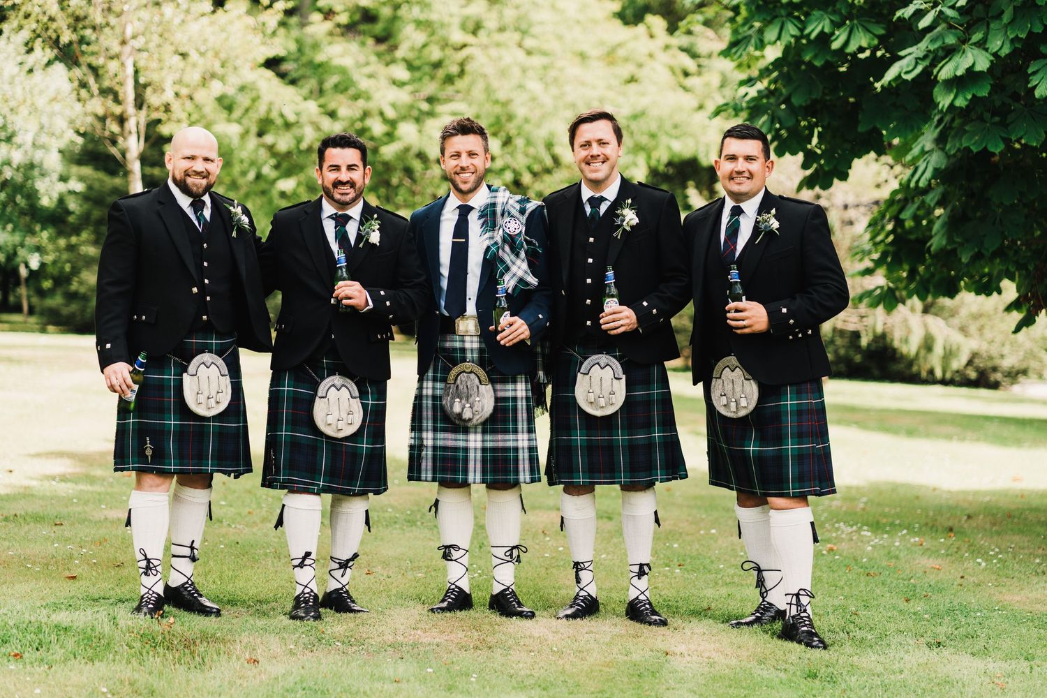 Five men in traditional Scottish kilts and formal wear stand in line on green lawn.