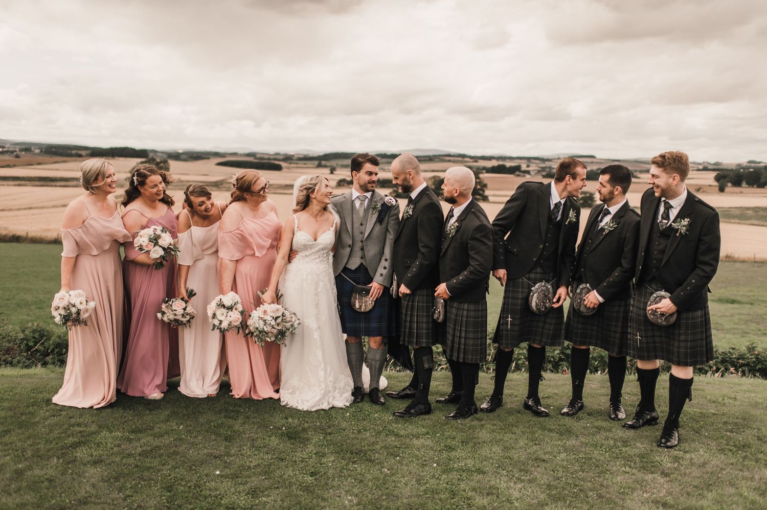 Wedding group photo with bridesmaids in blush gowns and groomsmen in traditional Scottish kilts against rural backdrop.