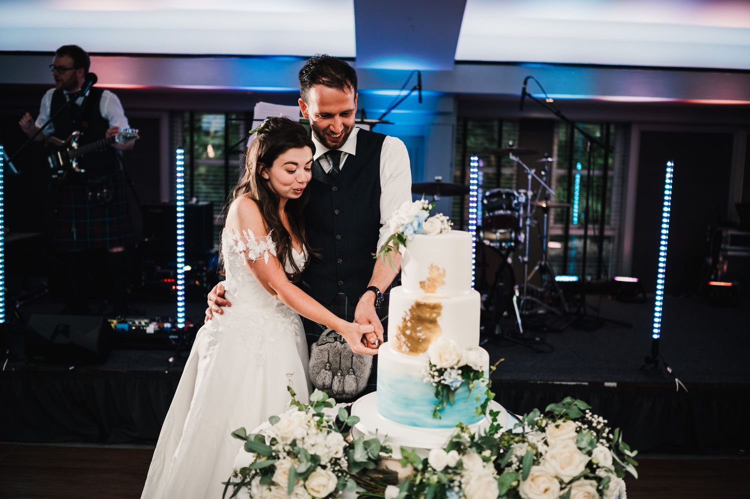 Newlyweds cut their beautiful white wedding cake together at indoor reception.