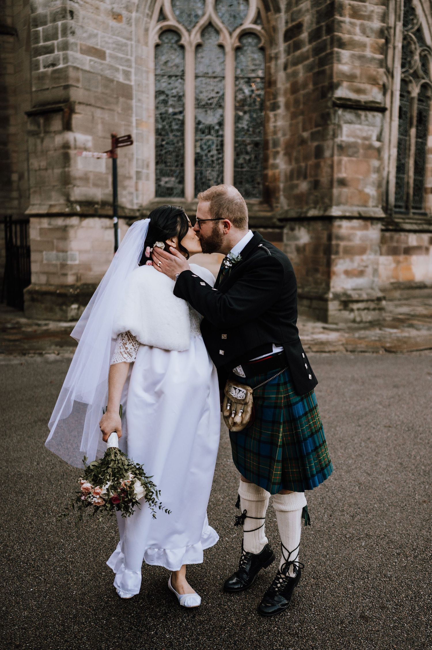 Romantic kiss between newlyweds in Scottish and traditional wedding attire by stone church.
