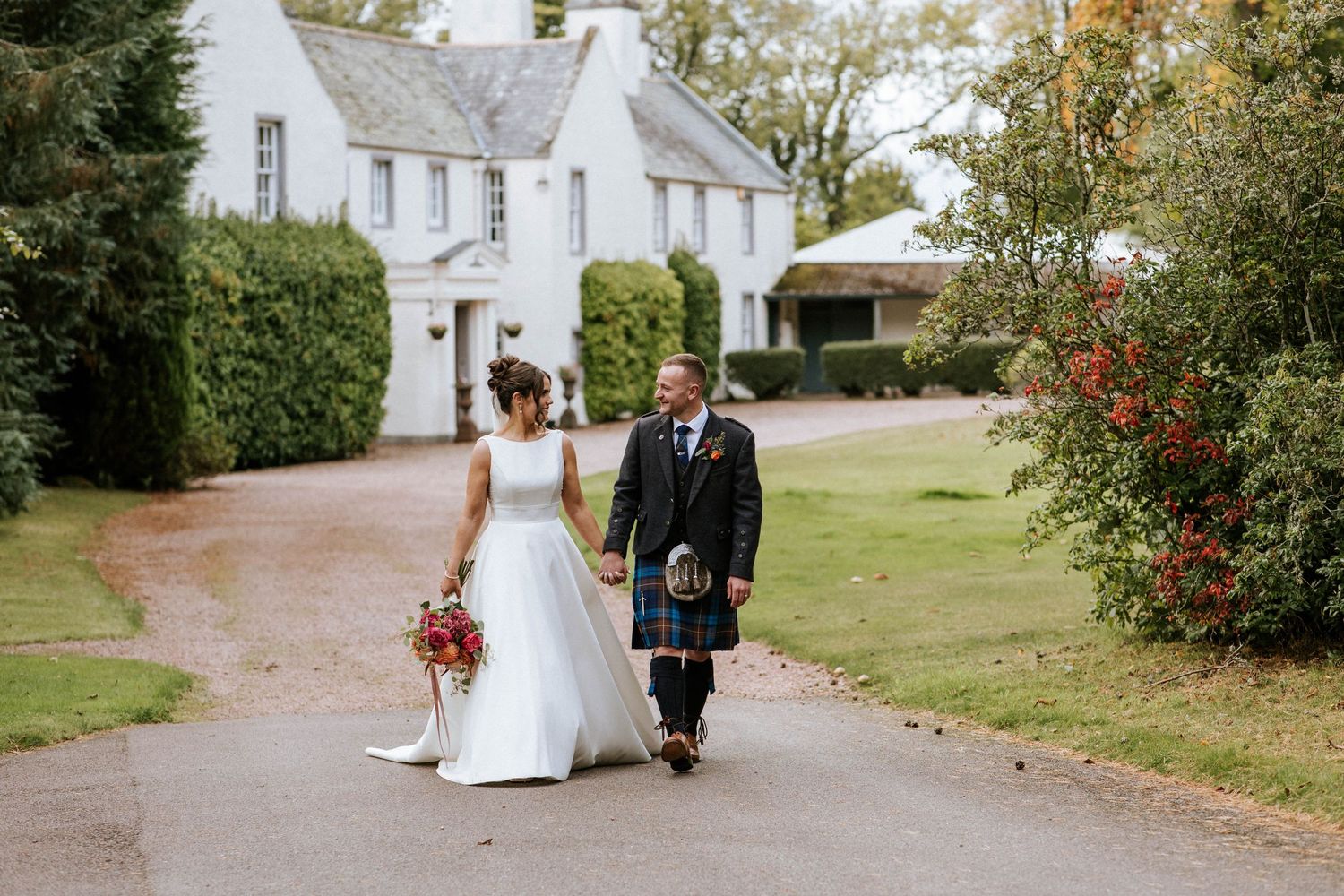 Wedding ceremony procession walking along rustic path beside white country buildings.