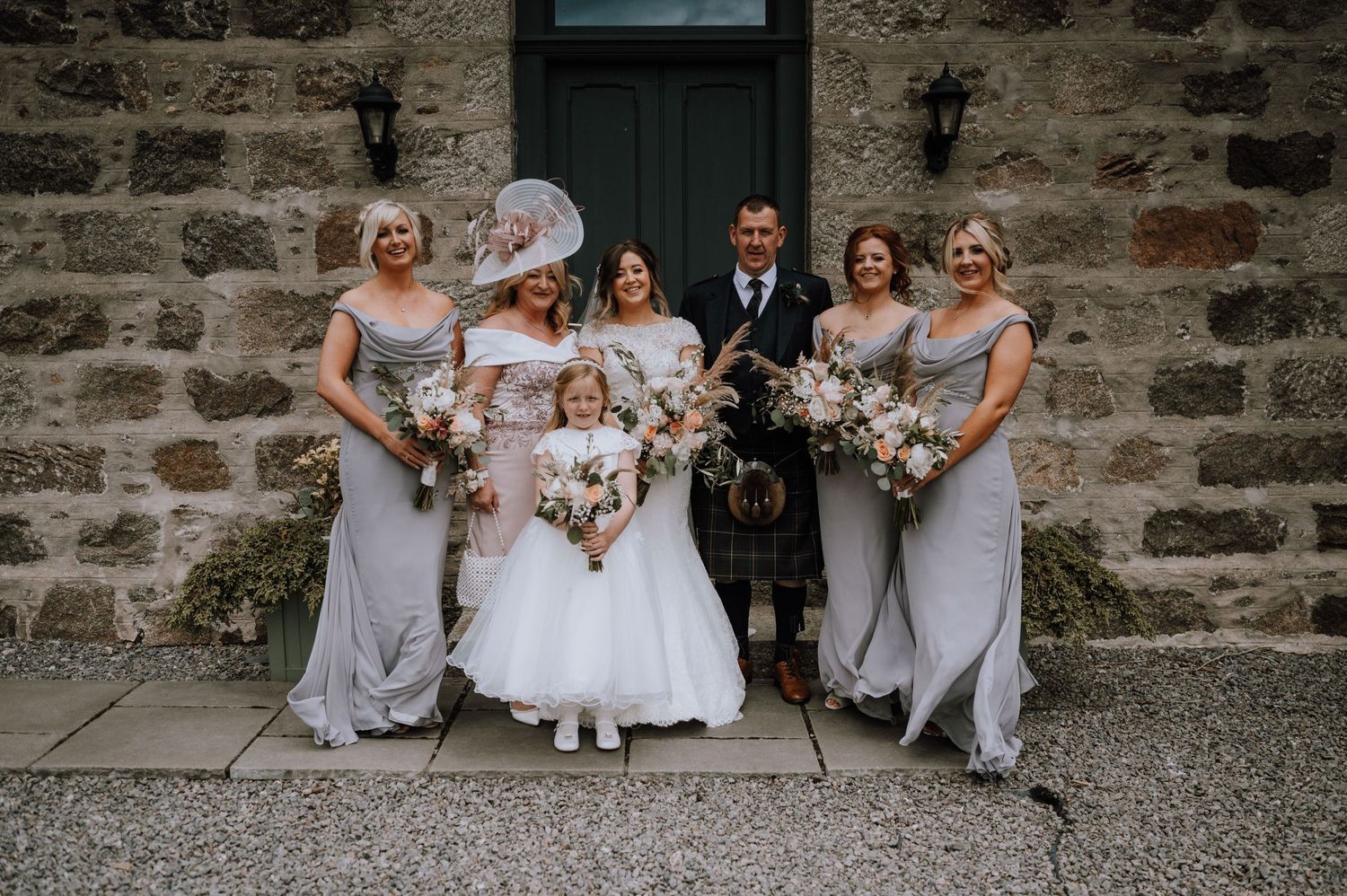 Wedding party poses together outside stone building with bridesmaids in lavender dresses and Scottish attire.