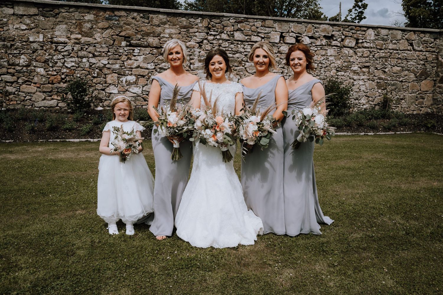 Bridal party in light gray dresses poses with bouquets against stone wall.