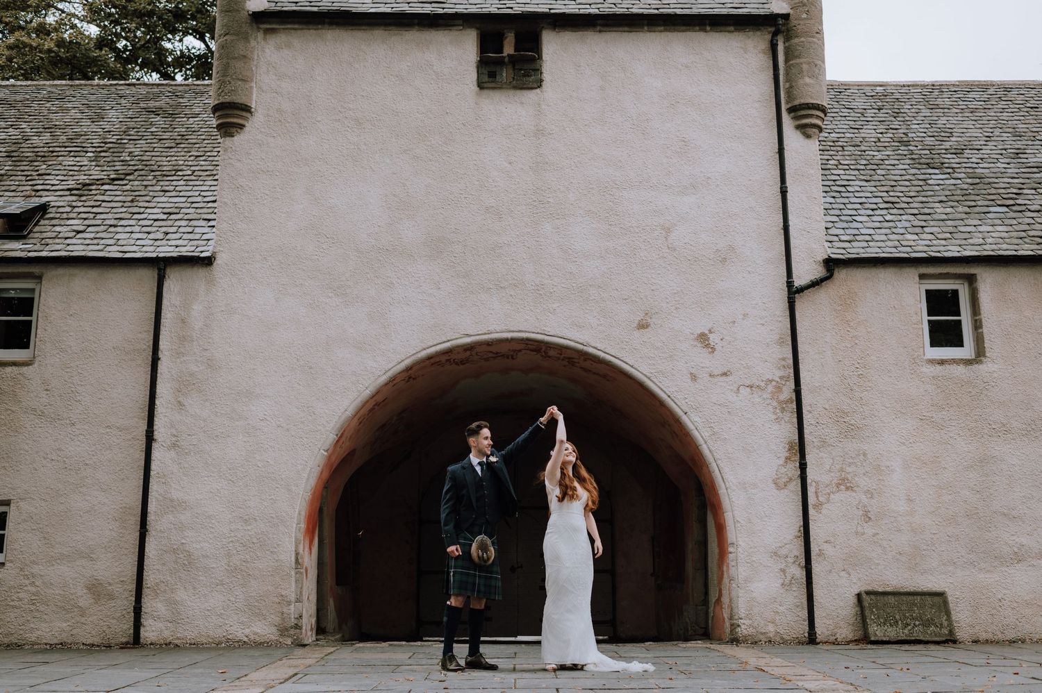Couple in wedding attire holding hands under stone archway of historic building.
